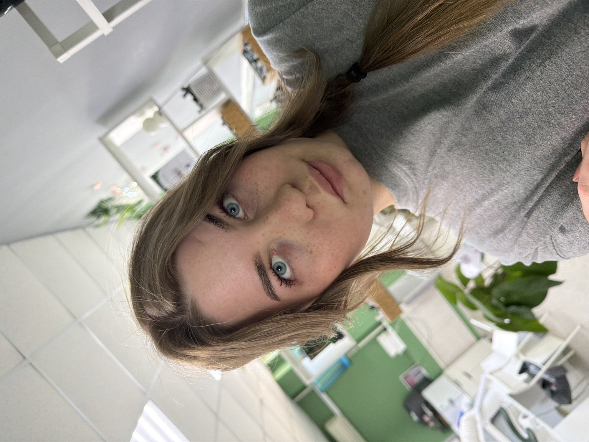 A medium close-up, slightly off-kilter, captures a woman with light-brown hair and fair skin, featuring freckles on her face. She is positioned in an indoor environment, possibly a commercial space like a salon or office. The background reveals a contemporary setting with white walls, modern furniture, and a green accent wall. The presence of chairs and equipment suggests the room's function. Her hair is partially pulled back, with the rest cascading around her face. She's wearing a grey, crew-neck shirt. The location is suggested as Chaikovsky, Russia based on visual context, which could be determined by geographical features or specific architectural styles, though these are not clearly visible.