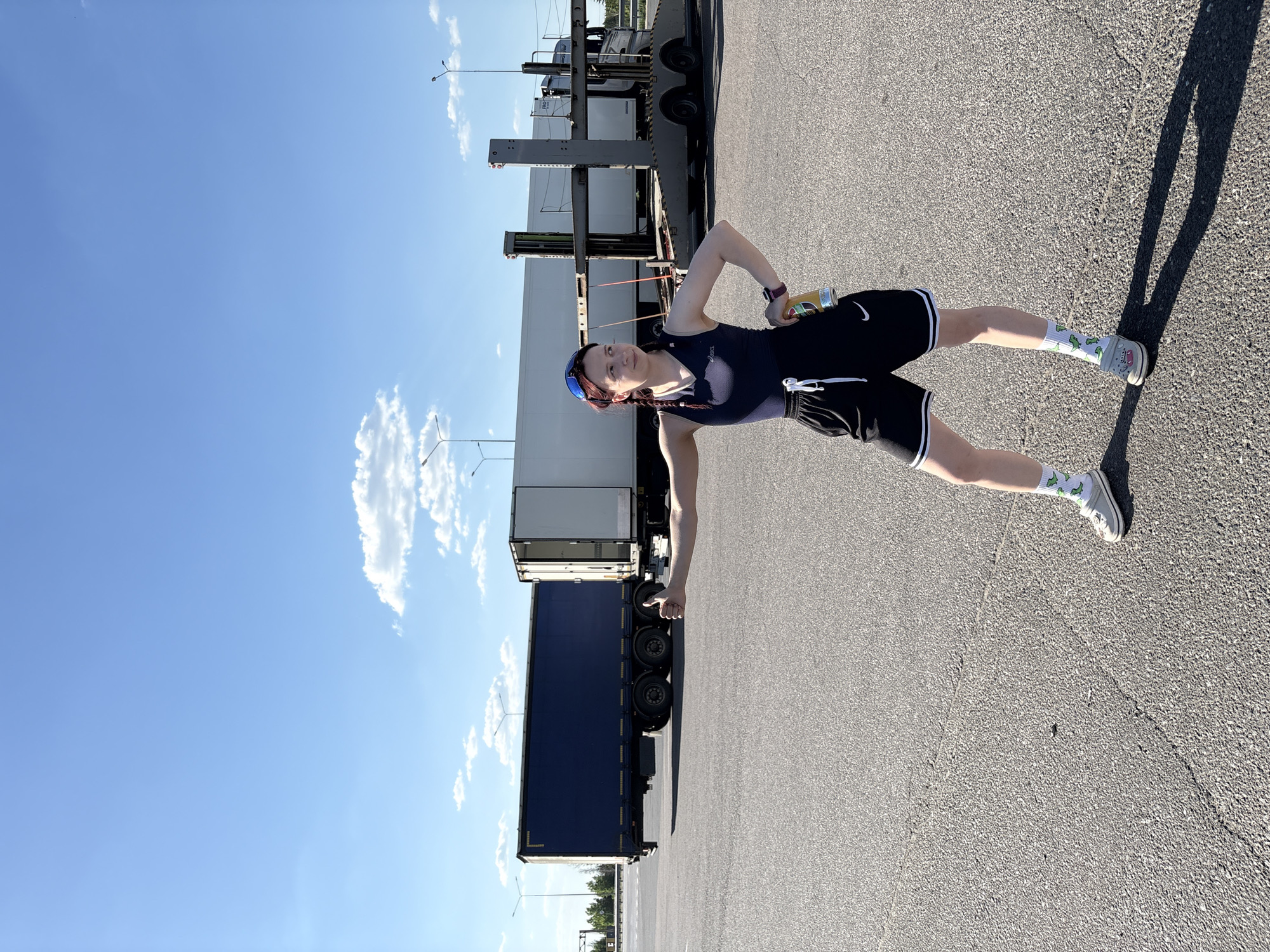 A woman stands on an asphalt surface with a large truck in the background. The woman is wearing a dark tank top, black shorts with white trim, white socks with a pattern, and grey sneakers. She has braided hair and is holding a can in her right hand. A large truck is parked behind her. The sky is blue with some clouds. The location is in Казань, Russia.