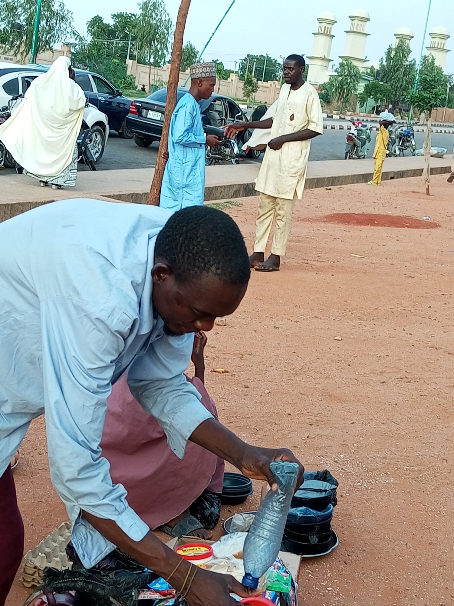 In Gombe, Nigeria, a man in a light blue long-sleeved shirt and trousers bends over a collection of goods spread out on the ground. He holds a partially filled plastic water bottle in his right hand and appears to be arranging items. Beside him, to his left, a person in a pink garment is seated, partially obscured. The ground is covered in reddish-brown sand, with scattered pebbles and some debris. In the background, several people are visible. One man in a cream-colored traditional outfit gestures with his left hand, facing another man in a blue traditional outfit and cap. Behind them, motorbikes are parked, and various vehicles, including cars, are on a paved road. The scene is set against a backdrop of trees and distinctive white domed structures, possibly part of a mosque or government building, characteristic of Gombe's urban landscape. The lighting suggests it is daytime.