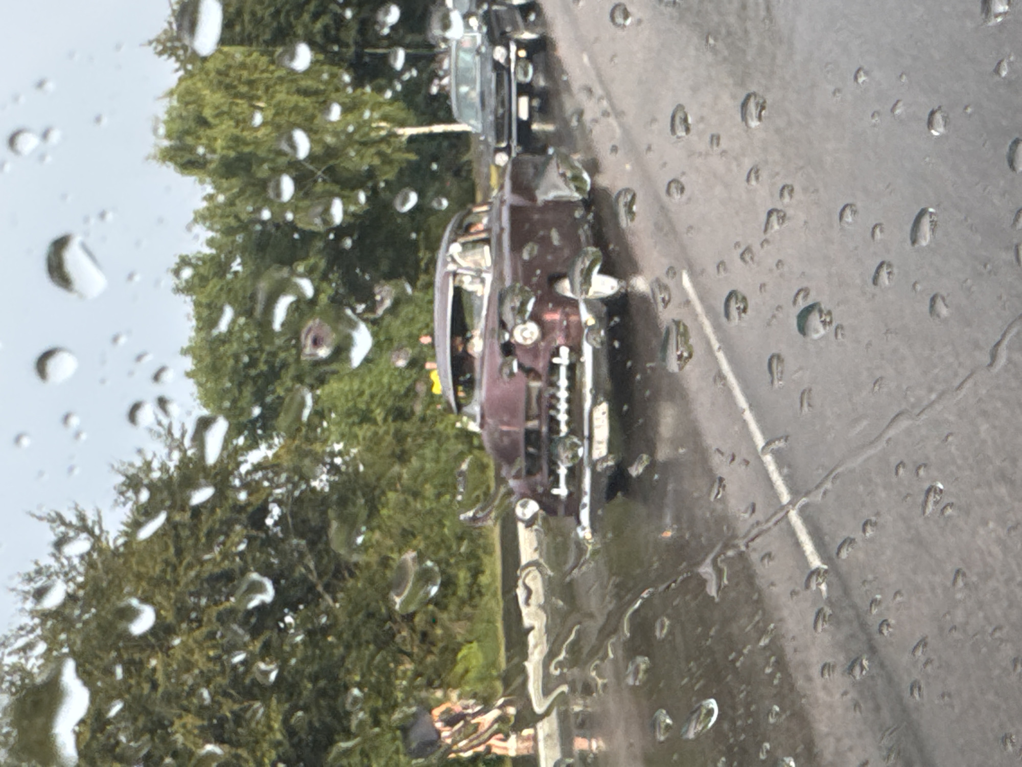 The image shows a vintage burgundy car driving on a wet street, with numerous raindrops obscuring the view. Trees and other cars are visible in the background, suggesting an outdoor setting, possibly in a city. The specific location in Falkenberg, Sweden, is not identifiable from the visual information provided in the image. There is a person walking on the side of the road holding an umbrella. The car appears to be in motion, and the overall atmosphere is one of a rainy day.