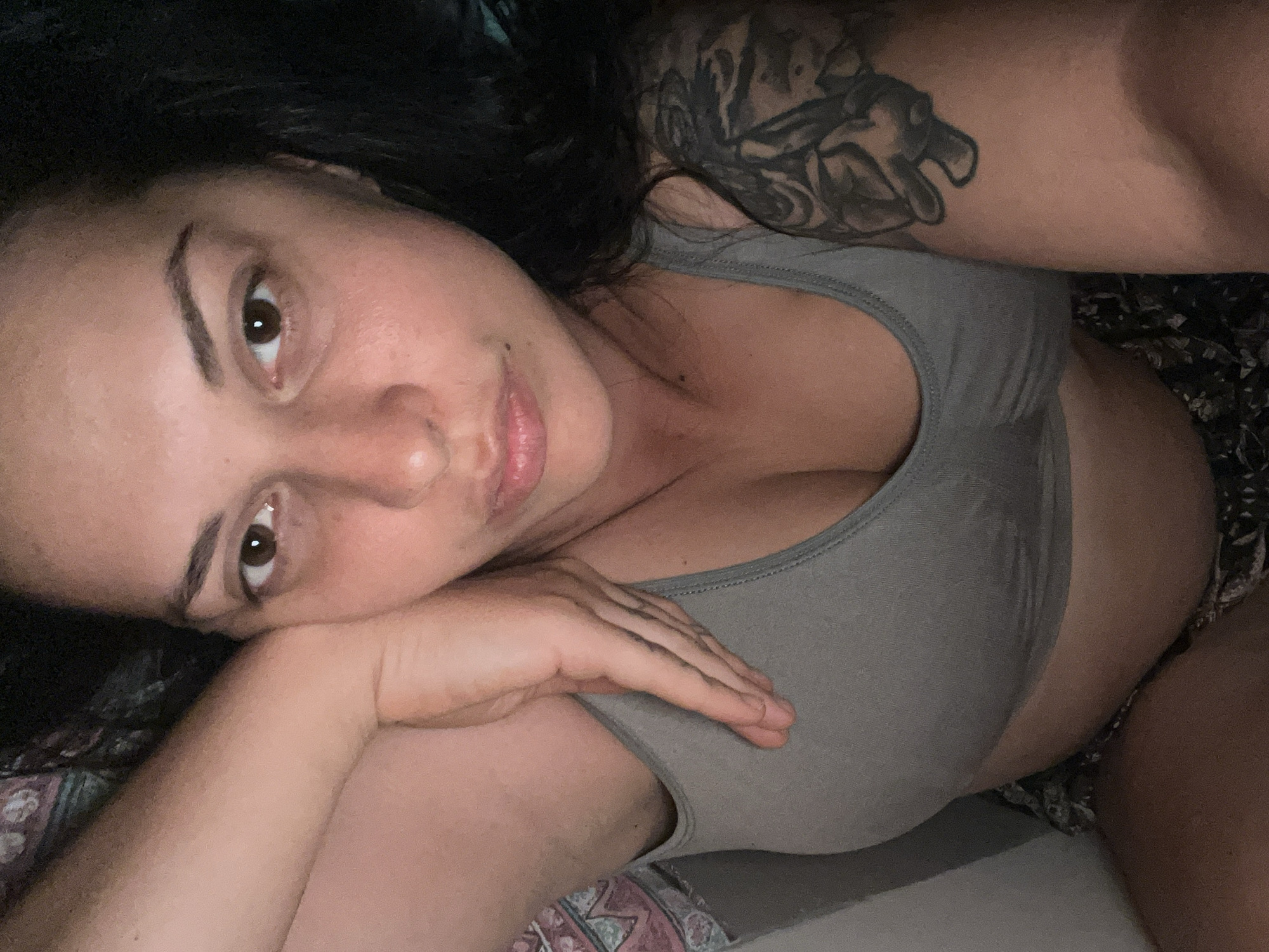The image is a close-up, high-angle selfie of a young woman lying down. She has dark hair and is looking directly at the camera with a neutral expression. Her right arm is bent, with her hand resting on her chin and cheek, partially obscuring her face. She is wearing a grey sports bra and appears to be in a bedroom or similar private setting. A tattoo is visible on her upper right arm. The lighting suggests it could be daytime or a well-lit indoor environment, with no specific weather cues. The setting does not offer any explicit visual clues to the location of Đà Nẵng, Vietnam.