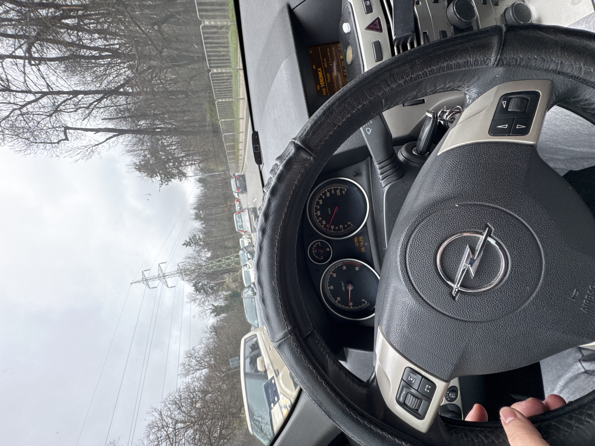 The image, taken from inside a car, shows the steering wheel with the Opel logo in the center. The speedometer and other gauges are visible behind the steering wheel. Through the windshield, there's a view of a road with several cars, likely in a traffic jam or queue. Trees without leaves line the road, and an electrical power pylon is also visible. The weather seems overcast. According to the car dashboard, it appears the radio station is “FMO VERONIKA”. The dashboard mileage reads 367626 km, and the driver is touching the wheel at the bottom right of the image. It is not possible to pinpoint the exact location to the city of Sofia with just this image.