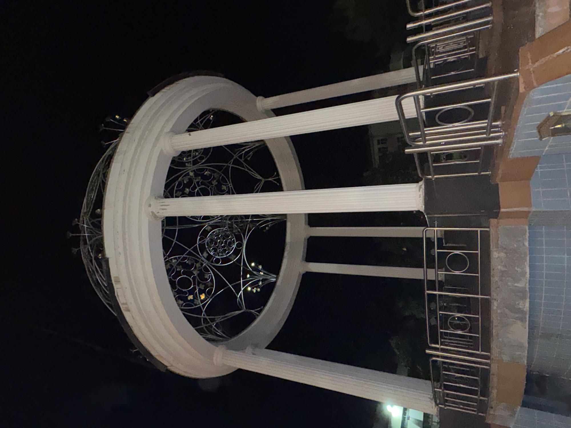 The image shows a low-angle shot of a circular, open-air pavilion at night in Botevgrad, Bulgaria. The pavilion has a white, tiered roof supported by fluted columns. The roof's edge is adorned with ornamental metalwork featuring circular and floral motifs, with small lights illuminating some of these designs. A metal railing with circular openings encloses the base of the pavilion, which sits atop a concrete structure. The foreground shows part of a tiled pool or fountain area. The background is dark, with only faint hints of trees and a distant building visible. The scene is illuminated by artificial lighting.