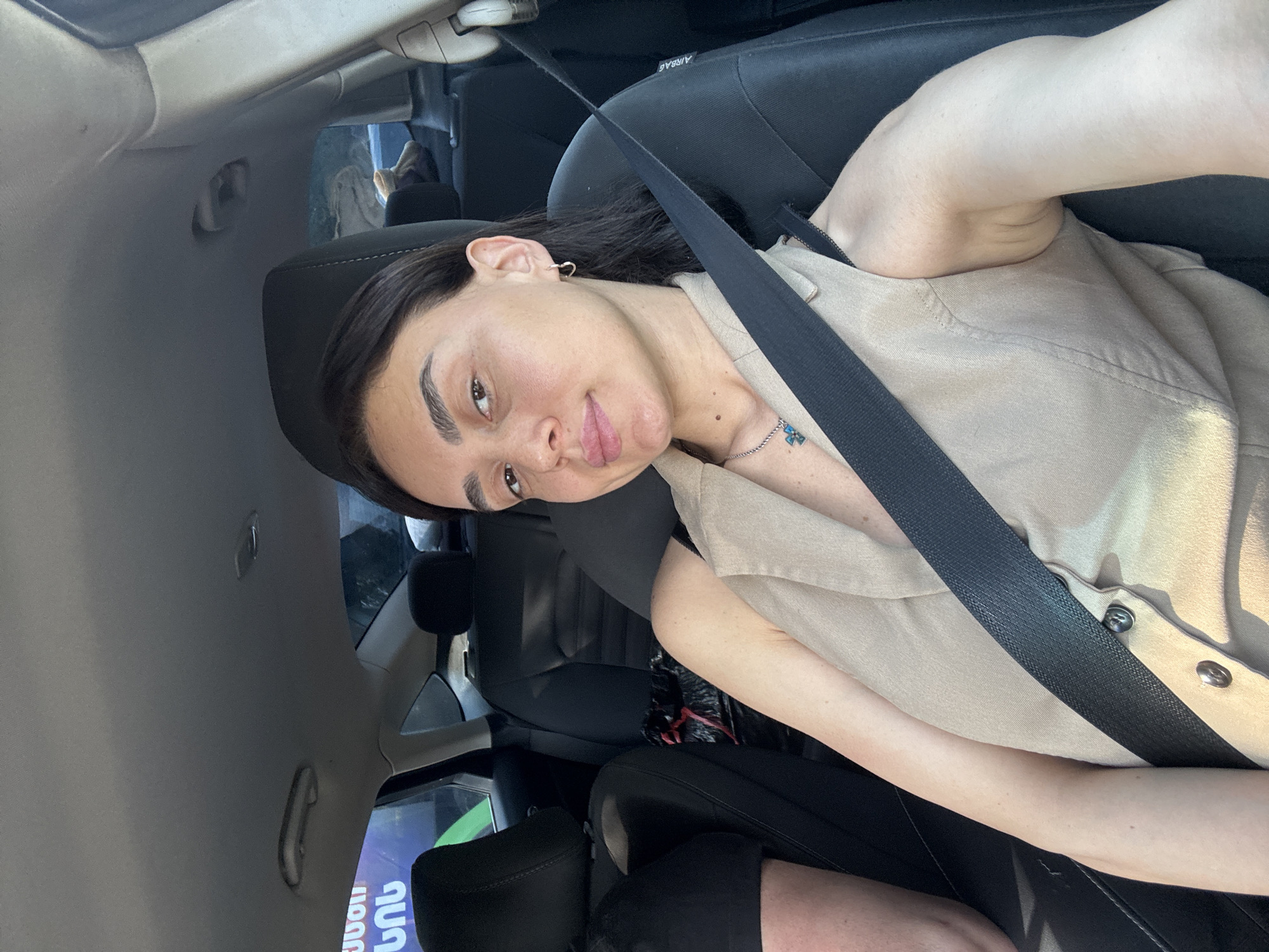 A woman with dark hair and light skin is sitting in the passenger seat of a car, looking at the camera. She is wearing a beige sleeveless top and a seatbelt. The interior of the car is visible, including the seats and the ceiling. Outside the car, there are trees and buildings. The image appears to be taken during the day, with natural light illuminating the scene. The text visible on a sign in the background is in Georgian, suggesting the location is Tbilisi, Georgia. The woman's facial expression is neutral, and she is wearing a subtle necklace.