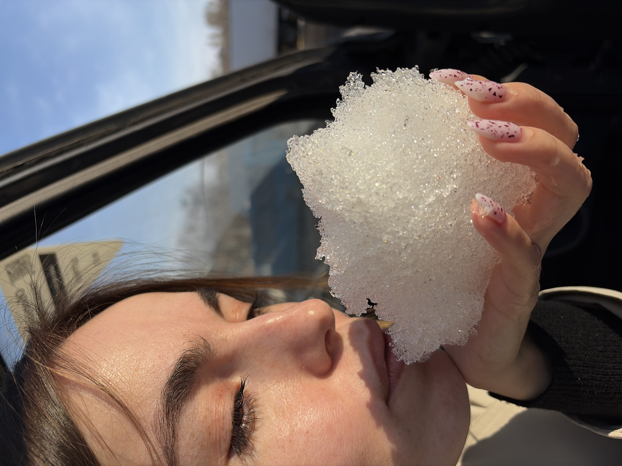 A person with long, dark hair is holding a ball of ice or snow up to their face. The person's eyes are closed, and they are wearing a beige jacket with a black undershirt. The person has pink and white nails. The background of the image shows a car window, a black car interior, and a blurry view of trees. The image has a bright and slightly overexposed quality.