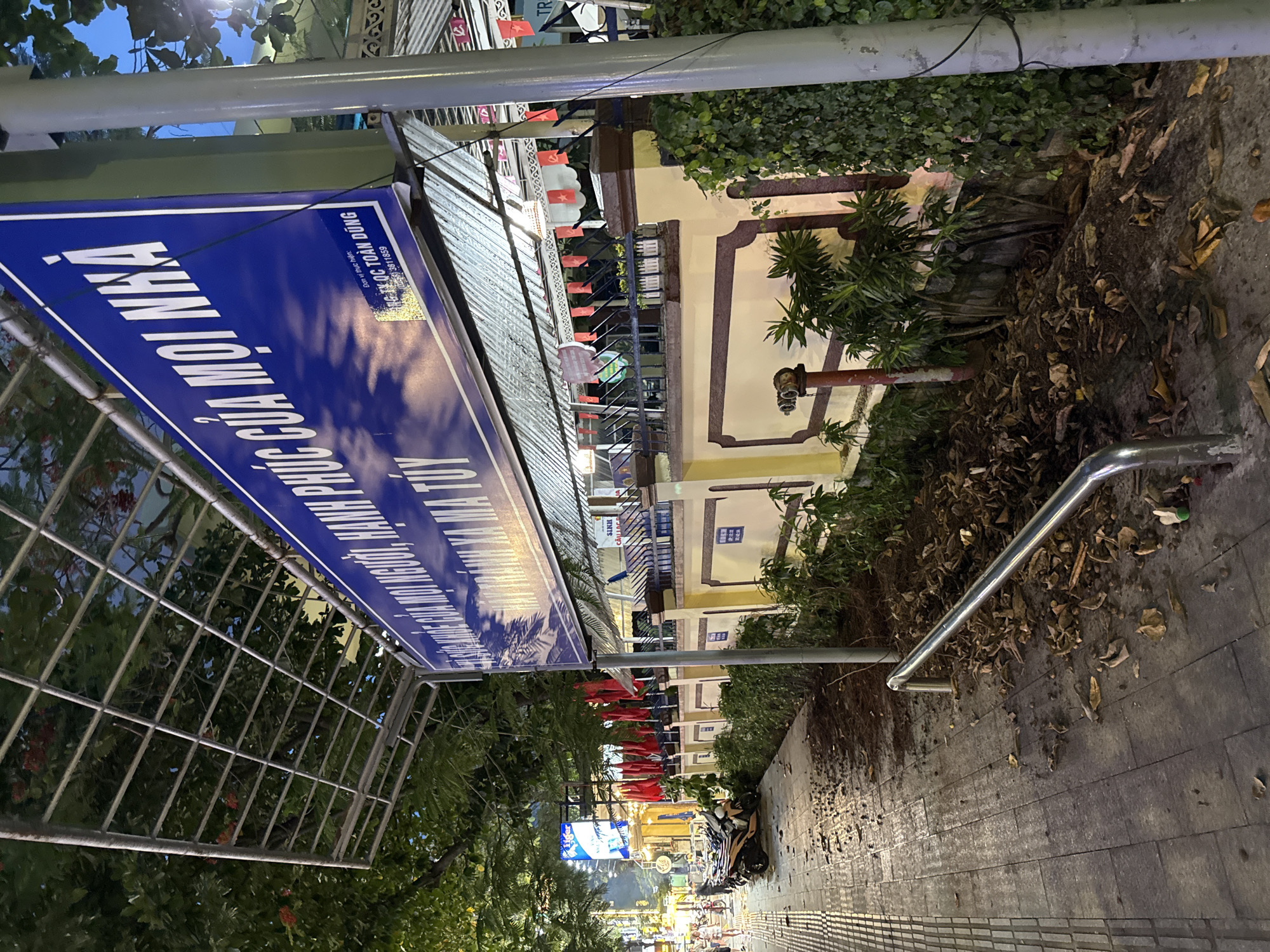 The image depicts a street scene in Nha Trang, Vietnam, likely in the evening. A large blue sign with white text, partially obscured by a metal grid structure, dominates the upper portion of the frame. The text on the sign appears to be in Vietnamese, with some English characters visible at the bottom. Below the sign, a building with a light-colored facade is decorated with red flags and banners. A row of potted plants and ivy adorns the front of the building. In the foreground, a sidewalk is littered with fallen leaves and debris. A metal railing runs along the edge of the sidewalk, and a fire hydrant is visible. Several motorcycles are parked on the sidewalk, and a few people can be seen walking in the background. The overall impression is one of a commercial or public area in Vietnam, with elements of daily life and street activity.