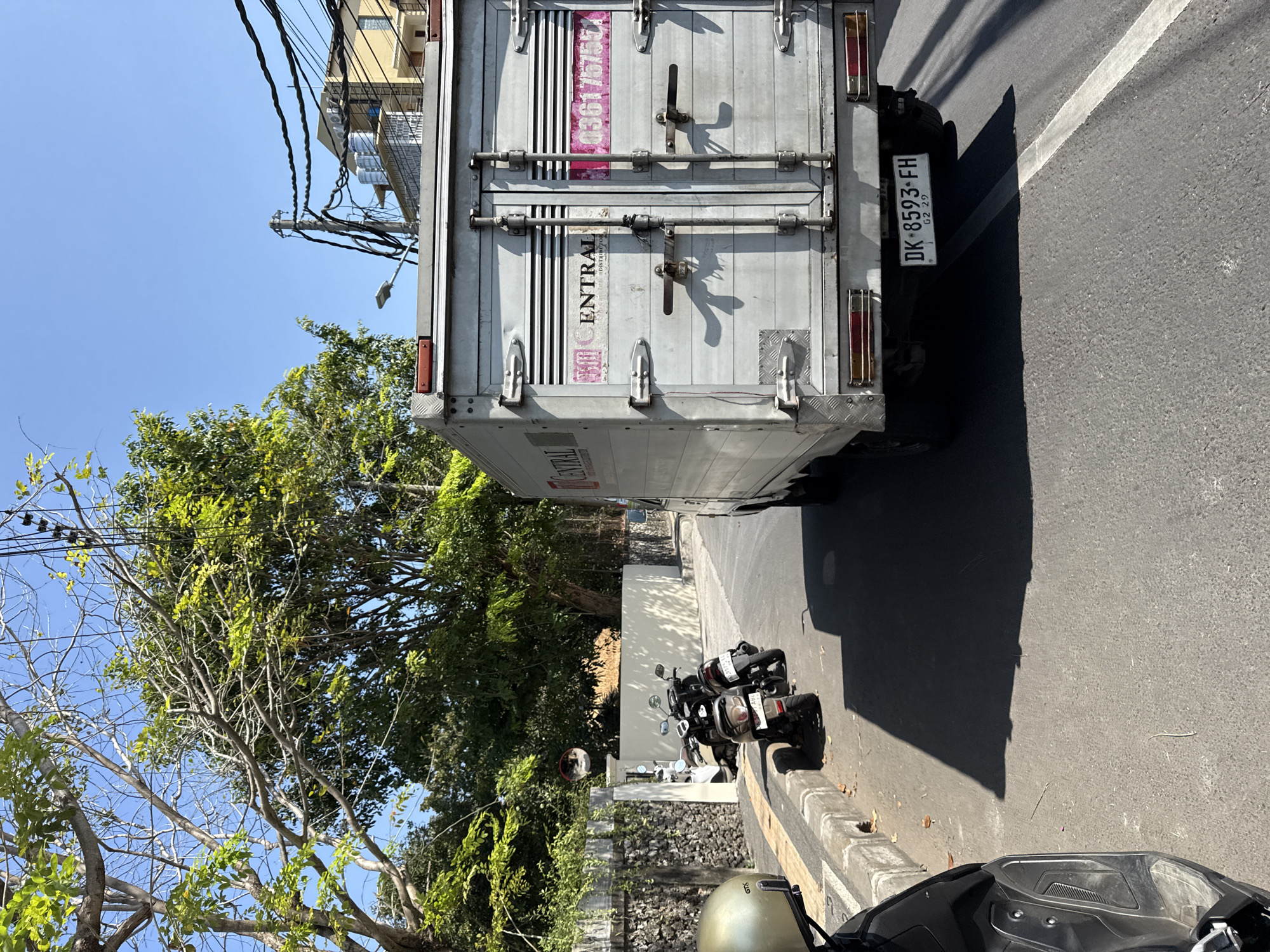 The image shows a street scene in Denpasar, Indonesia, on a sunny day. A white delivery truck with the brand "CENTRAL DISTRIBUTOR" is parked on the right side of the road, facing away from the camera. Its rear doors are closed, and a license plate reads "DK - 8593 - FH". To the left of the truck, several motorbikes are parked along the sidewalk. In the foreground, a portion of a black scooter is visible. The street is paved, and there is a concrete curb. Trees with lush green foliage line the left side of the street, casting shadows on the road. Power lines are visible overhead. In the background, there are buildings and more trees. A circular convex mirror is attached to a wall on the left side of the image.