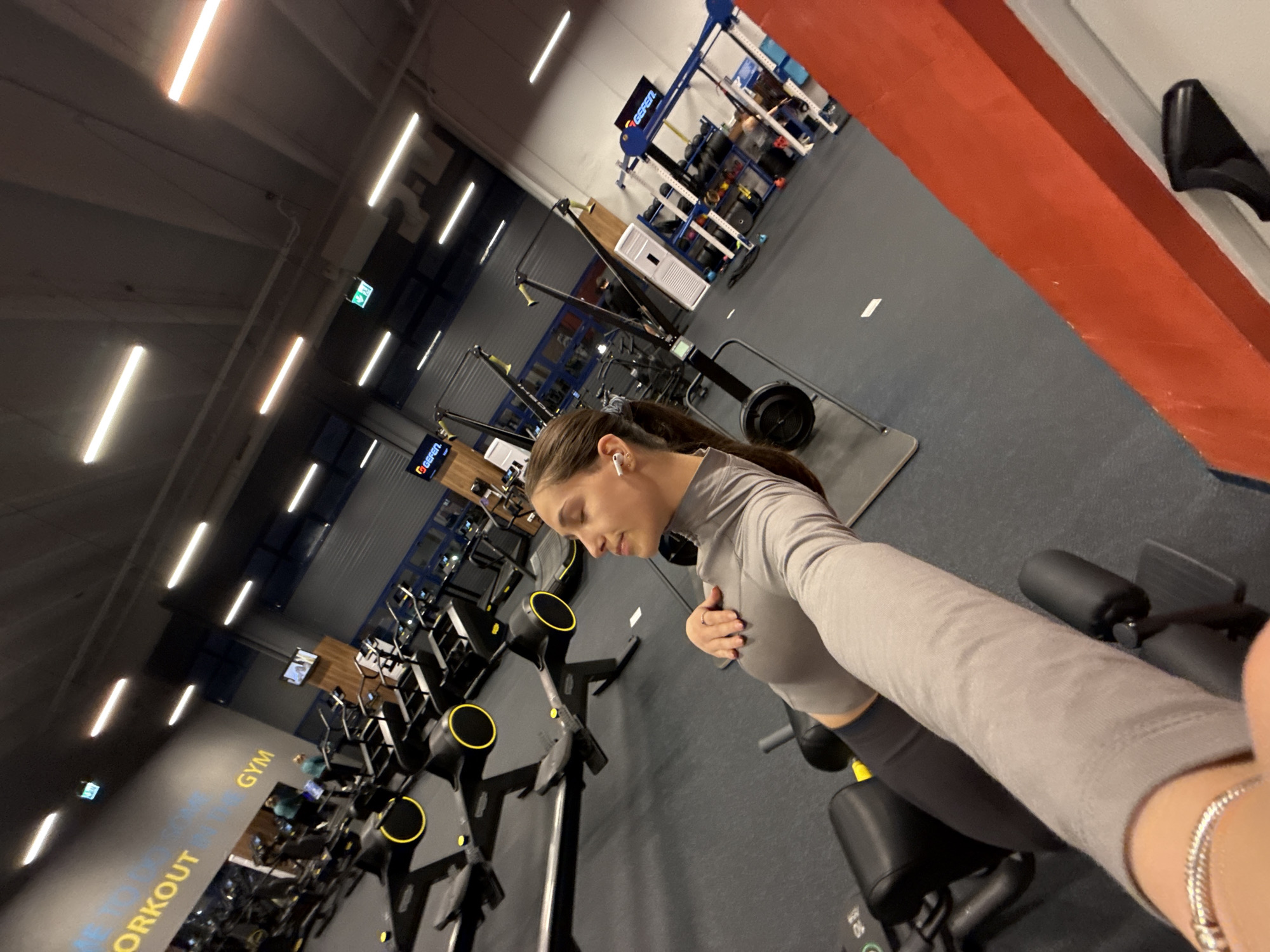 A young woman is taking a selfie in a well-lit gym. She is wearing a gray long-sleeved top and dark leggings, with a white earbud in her ear. Exercise equipment, including rowers and weight machines, are visible in the background. The gym appears to be located in Sofia, Bulgaria, based on the "WORKOUT FOR THE CITY" inscription on the wall. The location and specific equipment suggest a modern fitness facility.
