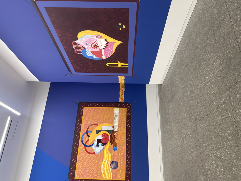 The image shows an interior with colorful abstract paintings. One painting hangs on the wall, featuring geometric shapes and lines, predominantly in shades of orange, yellow, and red. The other painting is located on the ceiling, in a dark red color with the same style of abstract geometric shapes. These abstract paintings are connected with a yellow and orange braided cord. The walls are painted in blue, white and gray.