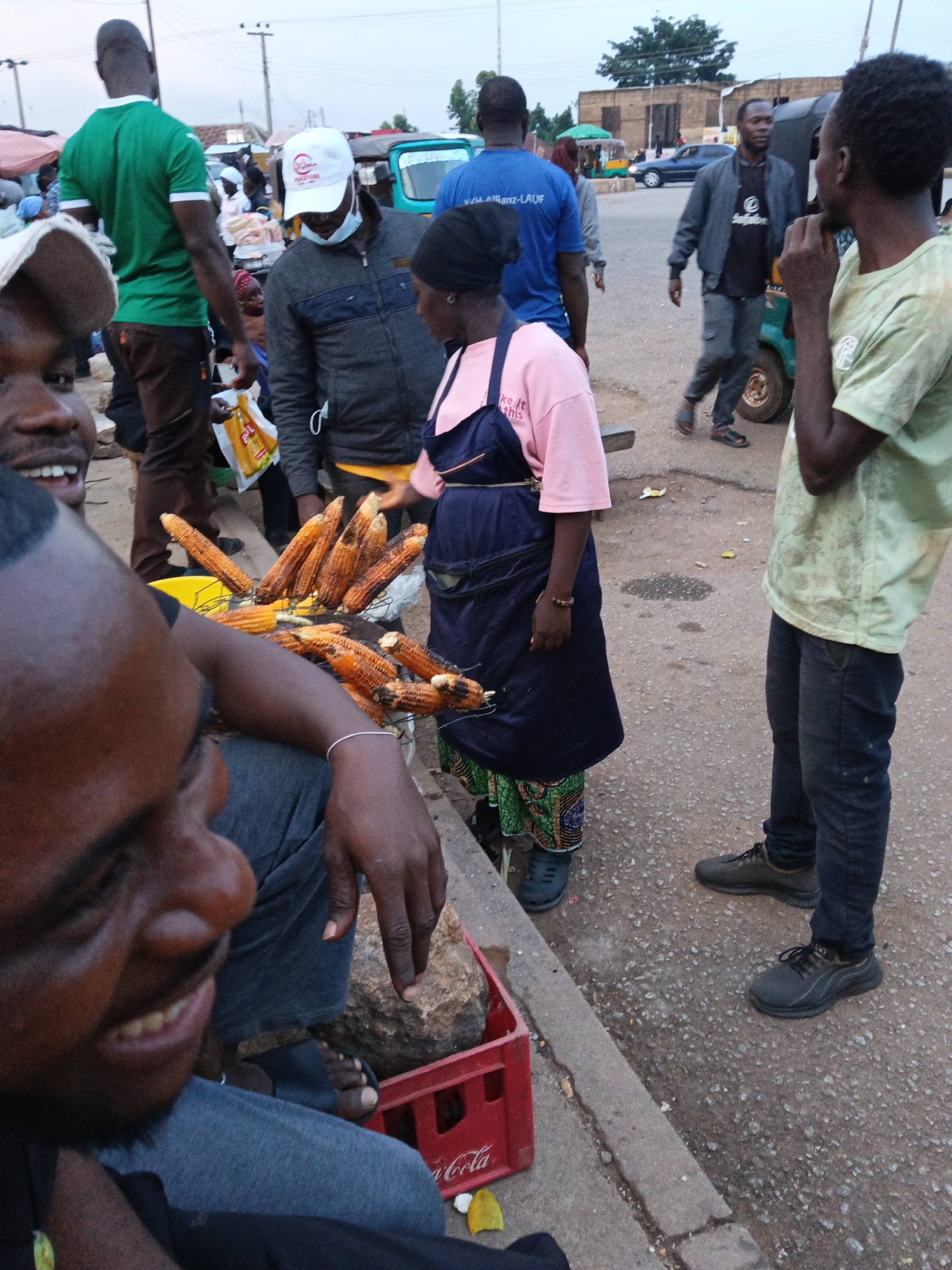 The image depicts an outdoor scene, likely a street or market, in an urban environment. Several individuals are present, some interacting with each other and with objects in the foreground.
In the immediate foreground, a man is partially visible, facing left, with his mouth open in a smile. He appears to be wearing grey trousers. To his right, a pile of roasted corn on the cob is displayed, resting on a metal grate. A red Coca-Cola crate is situated next to the corn.
Behind the corn, a person wearing a dark blue apron over a pink shirt and patterned skirt is seen from the chest down. They are facing right and appear to be interacting with the corn.
Further back, a man in a grey jacket and dark trousers is walking towards the right side of the frame. He is wearing a white cap and a light blue shirt. Next to him, another man in a blue t-shirt with text on the back is walking away from the camera.
To the far left, a man in a green t-shirt and brown trousers is standing and looking to his right. He is partially obscured by the foreground figure.
In the background, several vehicles, including what appears to be a tuk-tuk or auto-rickshaw, are visible. Buildings and trees form the backdrop of the scene, suggesting a commercial or residential area within a city. The lighting suggests it is daytime, possibly late afternoon. The overall impression is a bustling street scene with vendors and passersby.