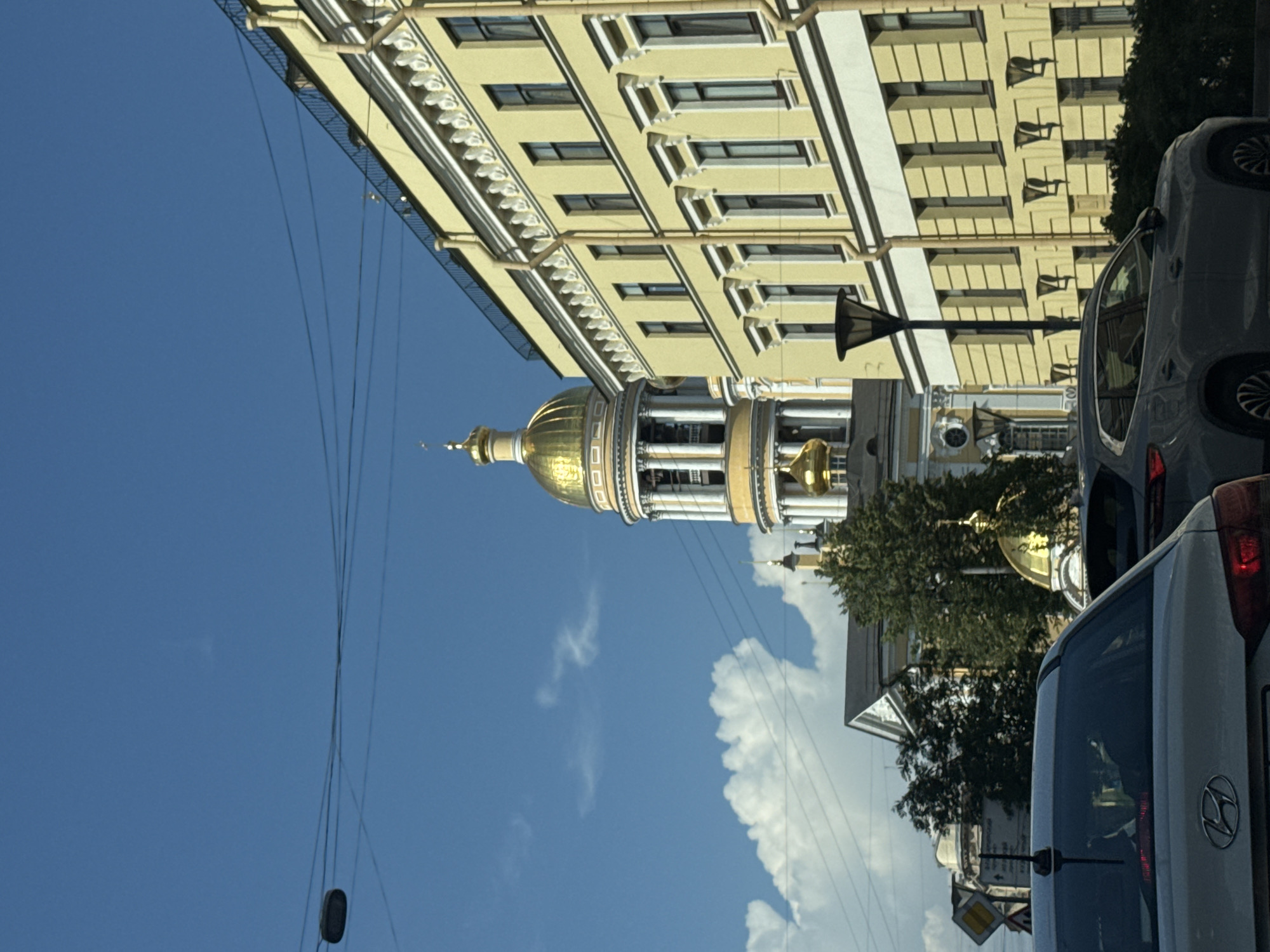 The image depicts a street scene in Saint Petersburg, Russia, featuring the golden dome of a church, likely St. Isaac's Cathedral given its prominent position and architectural style. The foreground shows the rear of a light-colored sedan and the side of a dark gray SUV, indicating traffic. A tall, modern street lamp stands to the right of the vehicles. Behind the cars, a large, pale yellow building with white trim and numerous dark windows dominates the right side of the frame. To the left, partially obscured by the yellow building and trees, is the multi-tiered structure of the church with its distinct golden dome and white columns. The sky is a clear blue with scattered white clouds. Electrical wires are visible crisscrossing the sky. The overall impression is a view of a historic urban environment with modern vehicles and infrastructure.