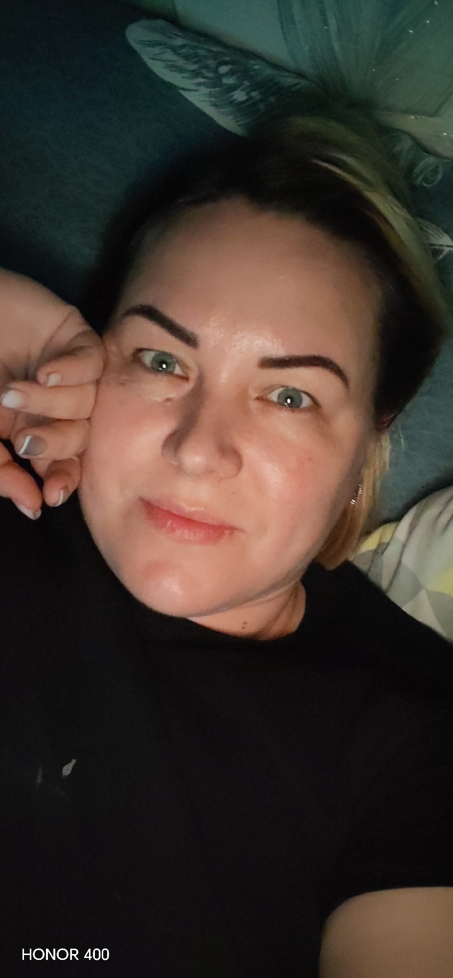 A selfie of a woman with fair skin, short blonde hair, and green eyes. She is lying down, with her left hand near her face. She is wearing a black shirt. The background includes a patterned blanket or pillow. The text "HONOR 400" is in the bottom left corner. The location is not specified.