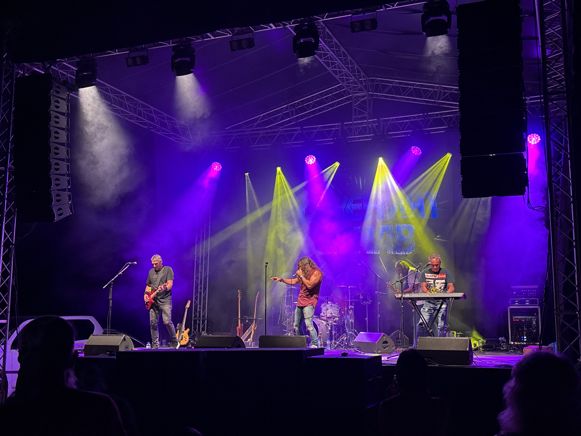 The image depicts a live music performance on an outdoor stage, likely in Tutrakan, Bulgaria, illuminated by purple and yellow stage lights. A band of at least four musicians is performing. In the foreground, a man in a dark shirt and ripped jeans is singing into a microphone, gesturing with his left hand. To his left, another man in a dark shirt and jeans plays a red electric guitar. Further to the right, a man wearing glasses and a dark t-shirt with a graphic print is seated at a keyboard. Behind the singers, a drum kit is visible. Several guitars are propped up on the stage, and large speaker cabinets are positioned on either side of the stage. The audience is partially visible in the foreground, with silhouetted figures watching the performance. The stage structure, including metal trusses and lighting equipment, is prominent above the performers. Smoke or fog is being used to enhance the lighting effects.