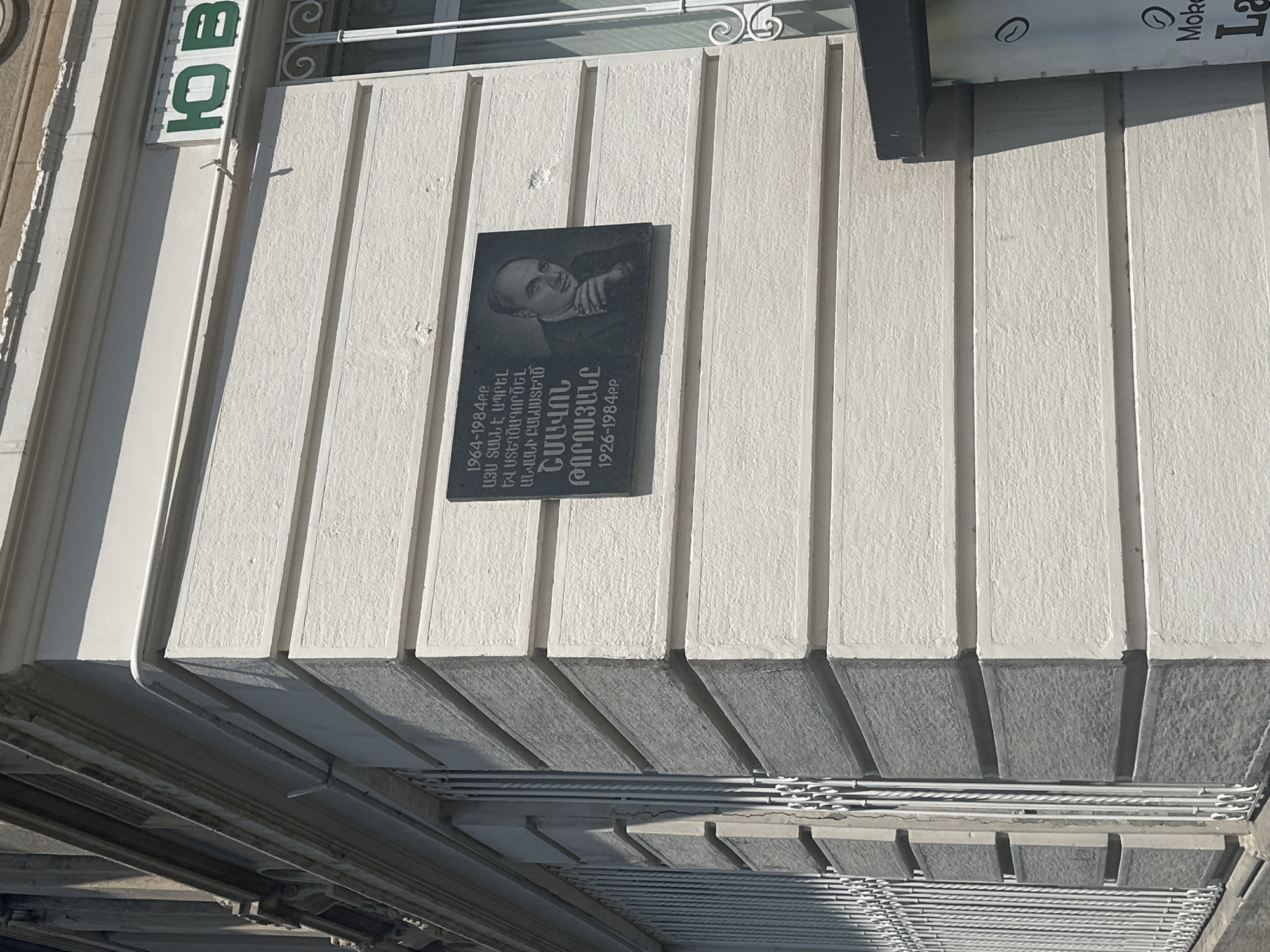 A plaque is mounted on a building in Gyumri, Armenia. The plaque features a portrait of a man and text in Armenian script. The building's facade is white with horizontal textured panels. To the left of the plaque, there are white metal bars covering windows. To the right, a window with ornate white metalwork is visible. Above the plaque, a sign with green letters on a white background reads "Ю В". A portion of a black awning and a sign with the text "Moko" and a partial "L" is visible at the bottom right. The overall scene depicts a section of an urban environment.