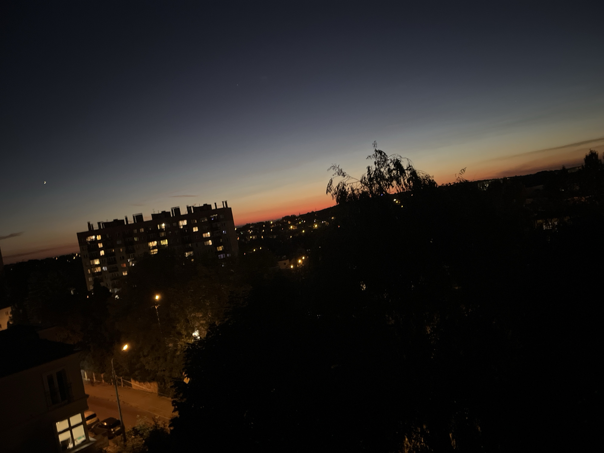 The image depicts a twilight sky over a city. A crescent moon is visible in the upper left quadrant. The sky transitions from a deep navy blue at the top to a gradient of dark orange and yellow near the horizon, suggesting the sun has recently set. In the mid-ground, a large, multi-story apartment building with many illuminated windows stands prominently on the left. Trees and other buildings with scattered lights dot the landscape, creating a bokeh effect in the distance. In the foreground, dark silhouettes of trees obscure much of the lower portion of the scene, with a hint of a street and a lamppost visible on the left. The overall impression is an urban landscape at dusk.