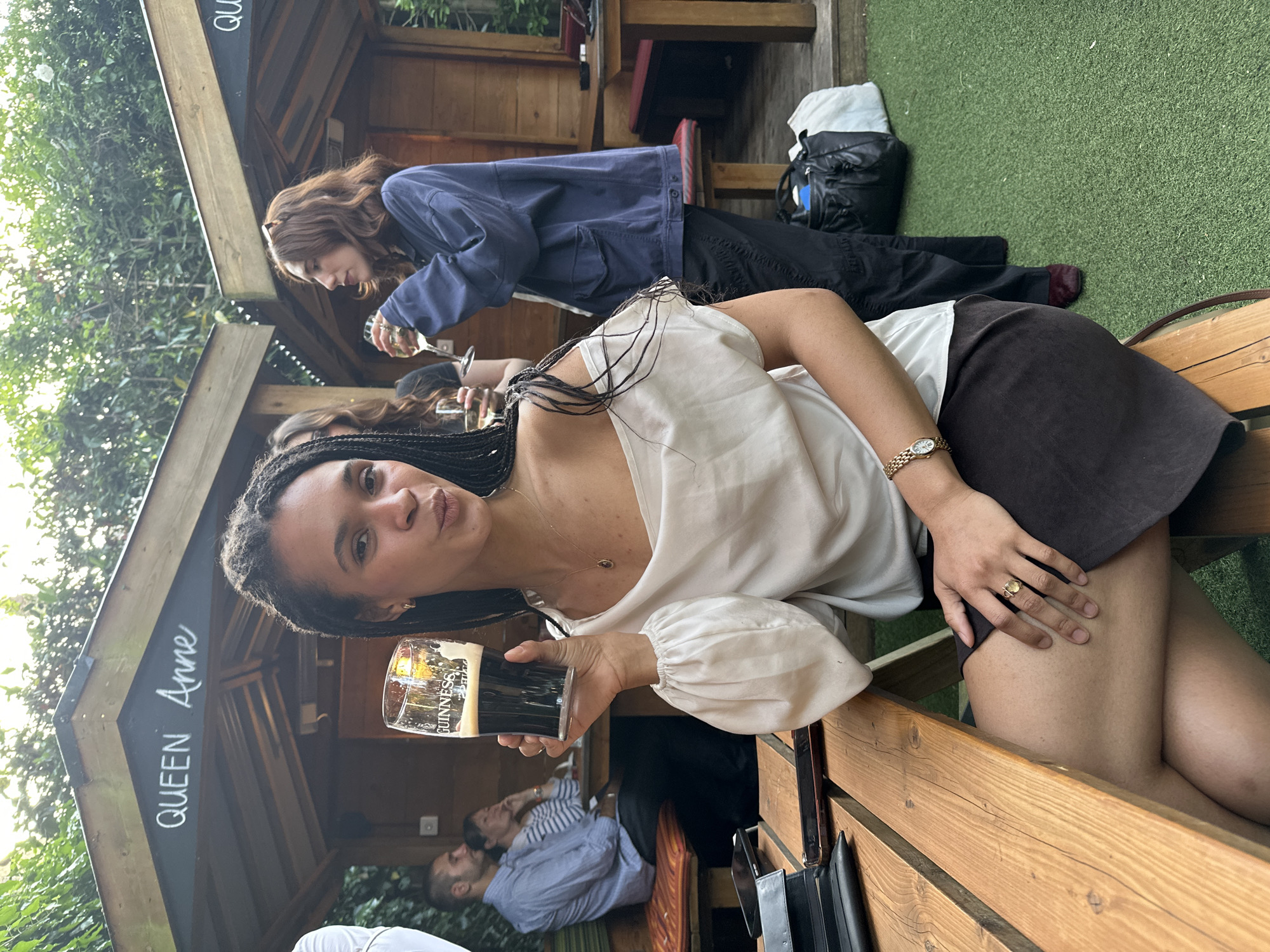 The photograph captures a scene at the Queen Anne pub in London, United Kingdom. A woman with dark hair braided with gold is prominently featured, holding a glass of Guinness beer. She is wearing a white blouse, brown skirt and a gold watch and ring. Other people can be seen in the background, including a woman with red hair wearing a blue jacket who is holding a wine glass, and two men sitting on a bench. The setting includes wooden benches and structures that are part of the pub's outdoor area, with glimpses of green foliage in the background. The name "QUEEN Anne" is written on the roof of the structure.