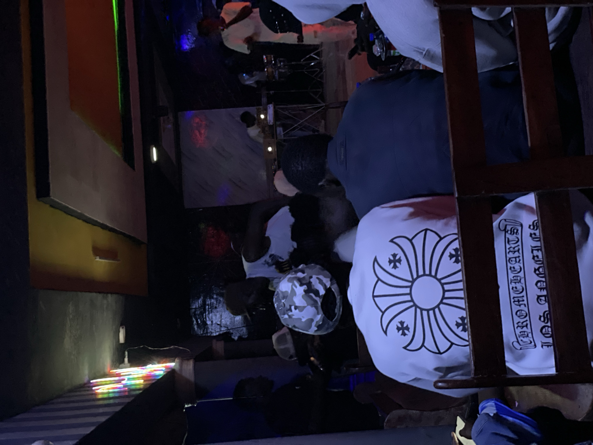 The image depicts an indoor gathering at night, possibly a bar or club, in Durumi, Nigeria. Several individuals are present, with a focus on those seated in the foreground and others interacting in the background. The lighting is dim, with colorful neon lights and spotlights creating a moody ambiance.
In the foreground, a person wearing a white t-shirt with a black Chrome Hearts logo and text "CHROME HEARTS LOS ANGELES" is seated facing away from the camera. To their right, another person in a dark blue jacket is also seated, with their back to the camera. To the left, partially visible, is a person in a light pink top and a person wearing a camouflage patterned hat.
In the background, several individuals are visible. One person in a white tank top appears to be interacting with someone else. Further back, a person is standing near a table with equipment, possibly a DJ or performer. Another person in a white shirt is standing on a platform, seemingly addressing the crowd. Musical instruments, including drums, are visible. The environment includes a ceiling with decorative panels and lighting fixtures, and walls with various textures and colors. The overall scene suggests a lively social event or entertainment venue.