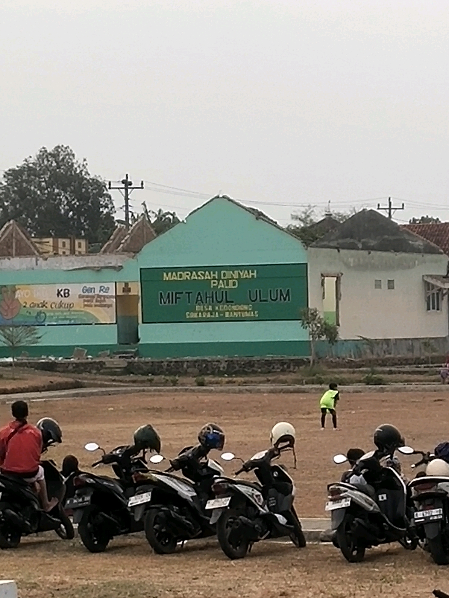 The image depicts the exterior of a building complex, likely a school or community center, in Desa Kedondong Kecamatan Sokaraja, Indonesia. The building features a prominent green facade with text that reads "MADRASAH DINIYAH PAUD MIFTAHUL ULUM DISA KEDONDONG SOQARAJA-BANYUMAS." The building's architecture is simple, with some sections appearing unfinished or in disrepair.
In the foreground, several parked motorcycles are visible. The motorcycles are of various models and colors, predominantly black. A few people are seen near the motorcycles. One individual, wearing a red jacket, is seated on a motorcycle. Another person in a bright green vest is visible in the middle distance, appearing to be participating in an outdoor activity on a dirt field in front of the buildings. The field itself is sparsely vegetated and appears to be a common area. The sky is overcast.