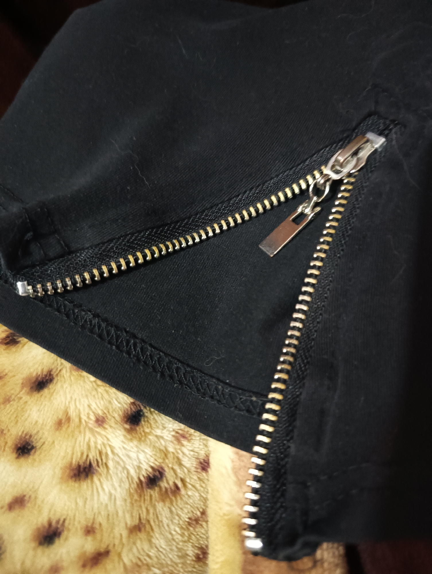 This close-up image captures a detail of black fabric, likely from a piece of clothing, featuring a metal zipper. The zipper has golden-toned teeth and a silver-colored slider and pull tab, and it runs diagonally across the frame. It appears partially open, revealing more of the black fabric underneath. The black fabric exhibits some minor lint and subtle creases. Below and to the left of the black garment, a distinct, textured fabric is visible. This second fabric is a warm yellow or beige with irregular dark brown or black spots, characteristic of a faux fur or leopard print material. The lighting emphasizes the metallic elements of the zipper and the textures of both fabrics. There are no people visible, and no specific activity is taking place. No text is discernible in the image. The scene offers a detailed view of these textile components, originating from Biliaivka, Ukraine.