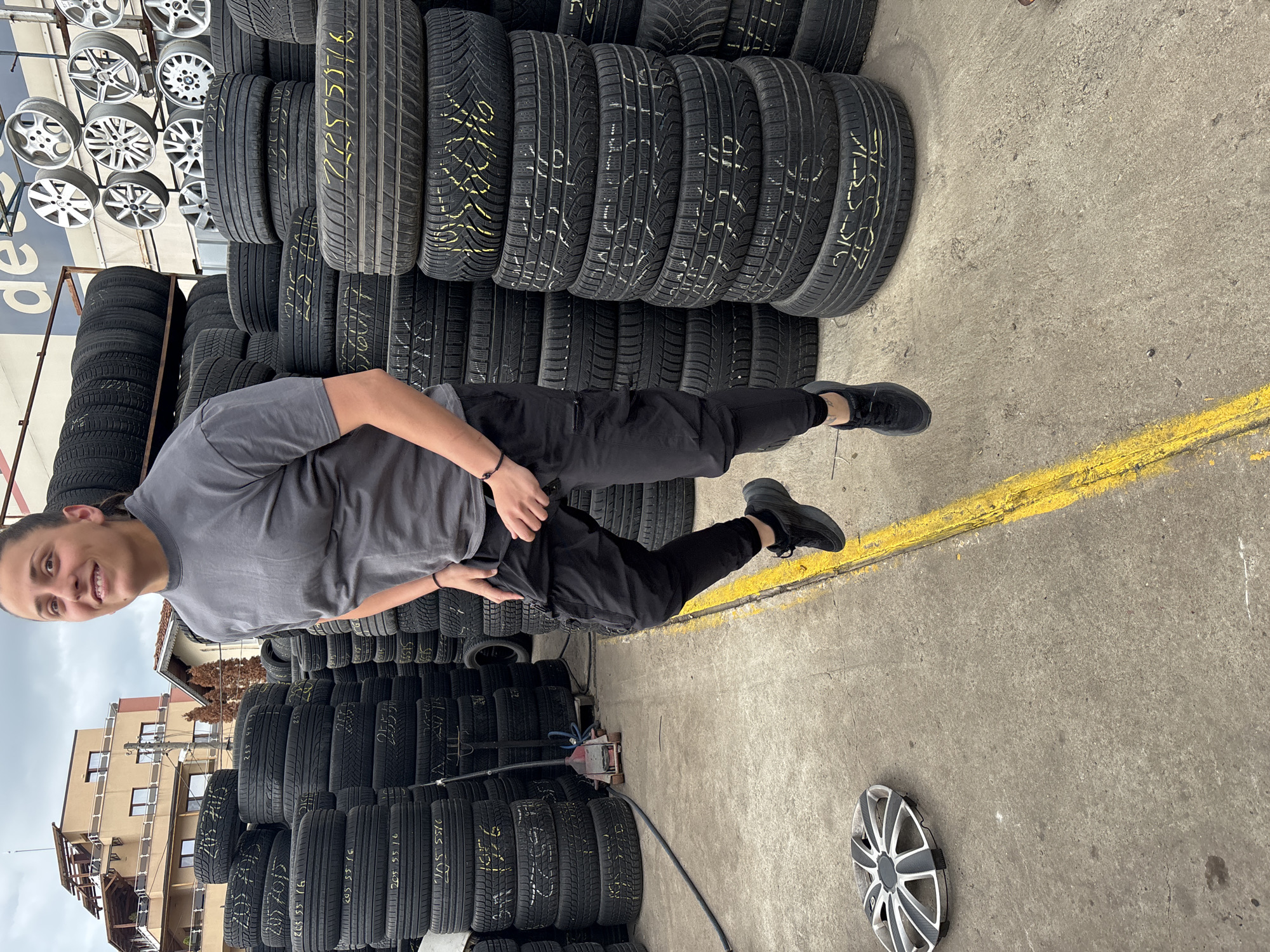 The image captures a person standing amidst stacks of tires. The setting appears to be a tire storage area. In the foreground, a person is positioned, facing the camera with a slight smile. They are clad in a grey t-shirt and black pants, with black footwear. The individual is standing beside a stack of tires that are labeled with yellow lettering. To the person's left, stacks of wheels are visible. Behind the stacks of tires and partially obscured by them, a building can be seen. The foreground is a concrete surface marked with a yellow line. A single wheel cover lies discarded on the surface. The general atmosphere suggests an industrial or commercial setting, likely related to automobile maintenance or sales. Based on available details, the location could possibly be in Sofia, Bulgaria.