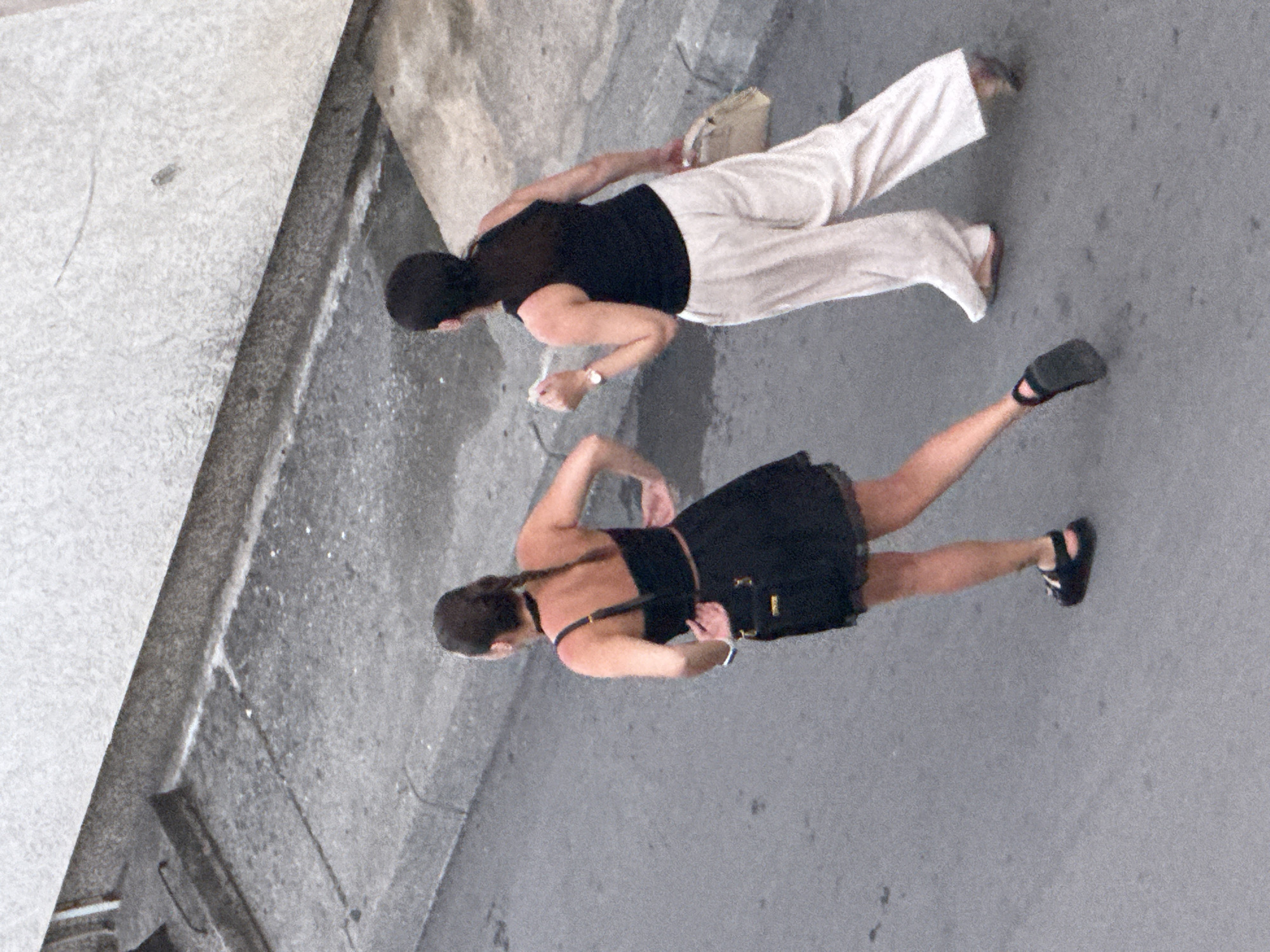 Two women walk on a paved road in Budva, Montenegro. The woman on the left wears a black dress and black sandals. The woman on the right wears white pants and a black tank top. She is holding a clutch bag in her left hand and a cigarette in her right hand. The road is bordered by a concrete sidewalk and a building.