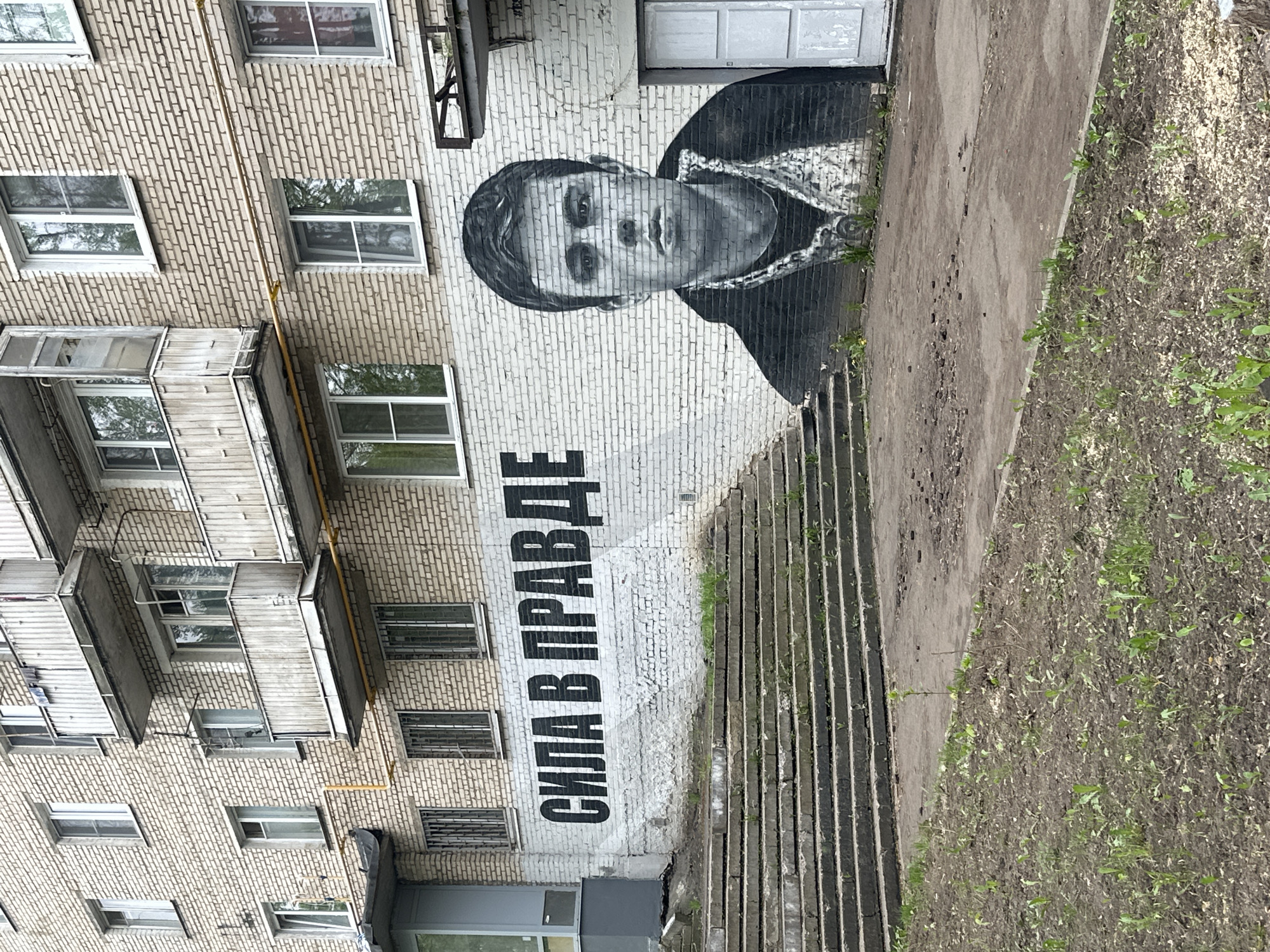 A photograph captures a mural on the side of a multi-story brick building. The mural depicts a black and white portrait of a person and, below the portrait, text written in the Russian language. The building has numerous windows and balconies. Adjacent to the building is a walkway and some grassy areas with vegetation. The scene is located in Krasnogorsk, Russia.