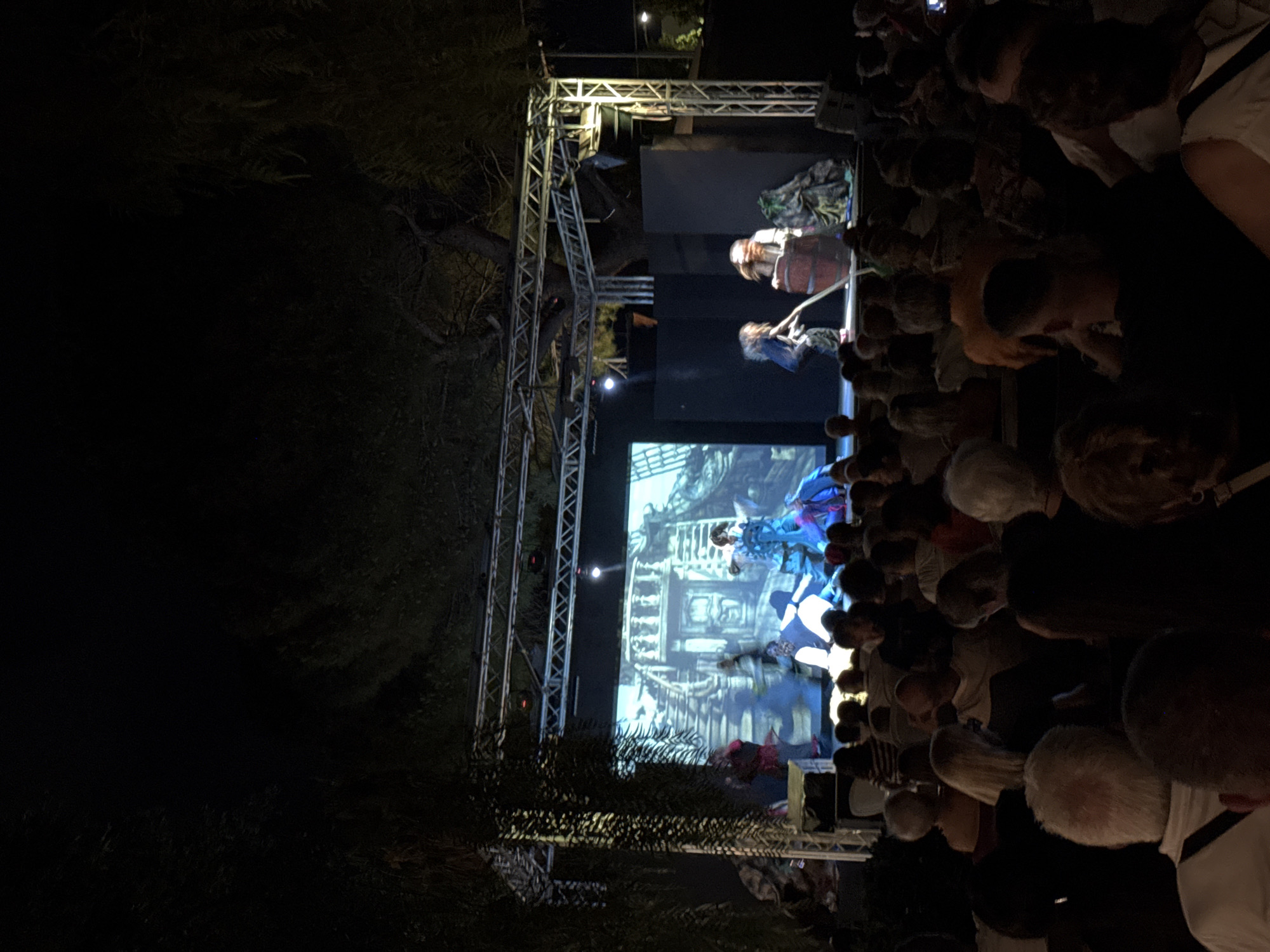 An outdoor evening performance is taking place on a stage in Isola delle Femmine, Italy. A large screen behind the stage displays a digital backdrop depicting a spooky, dilapidated building with stairs. On the stage, several performers in costumes are enacting a scene. One performer in a blue dress is near the center, while others in darker clothing are engaged in various actions. To the right, a large barrel is visible, and a performer is interacting with it. The stage is illuminated by stage lighting, and a metal truss structure supports the lighting and sound equipment. In front of the stage, an audience is seated in rows, facing the performance. The surrounding environment is dark, with trees visible in the foreground and background, suggesting the performance is taking place outdoors, possibly in a park or a public square. The image captures a moment during the performance, with the audience engrossed in the show.