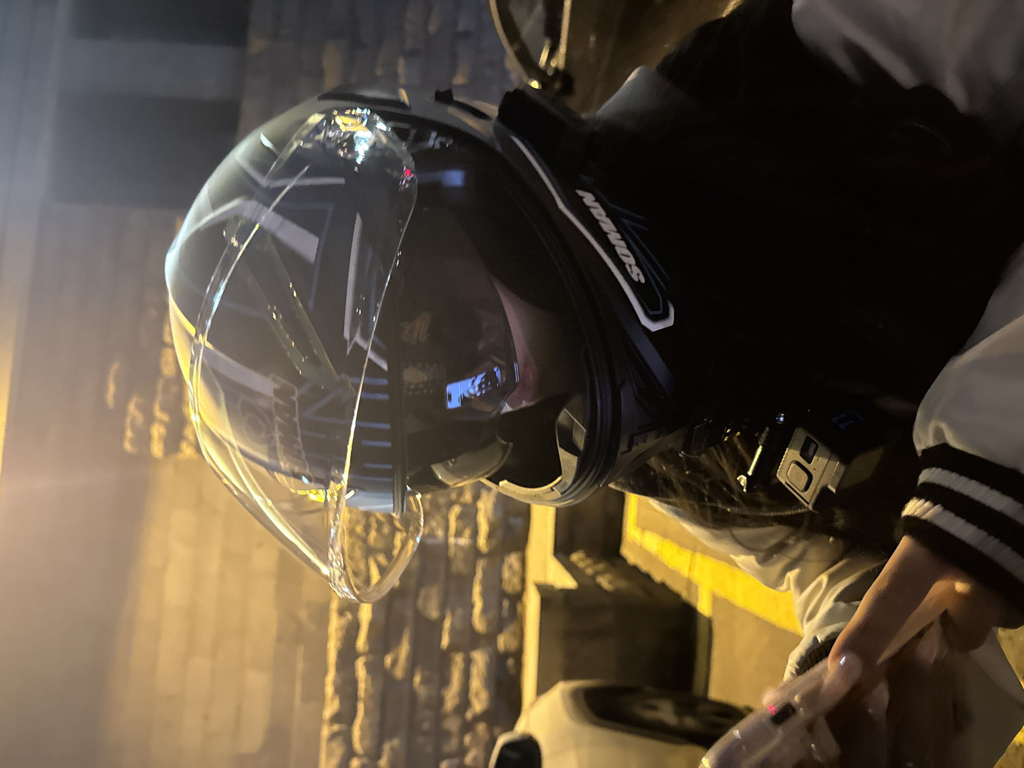 The image depicts a person wearing a full-face motorcycle helmet, viewed from a side profile at night in an urban setting, likely Yerevan, Armenia. The helmet is dark with white and grey graphic accents, and a clear visor partially obscures the wearer's face while reflecting warm, yellow-orange light from the environment. An action camera, possibly a GoPro, is mounted on the right side of the helmet. The word "SOMAN" is visible on a strap or side panel of the helmet.
The person is wearing a dark-colored jacket, possibly black, with white stripes on the sleeves, suggesting a sporty or varsity style. In the lower right corner, a hand is visible, featuring long, manicured fingernails and a ring on one finger.
The background is dimly lit by streetlights, casting a warm, golden glow on a textured wall, which appears to be made of stone or brick. Parts of a light-colored vehicle are faintly visible at the bottom of the frame, suggesting the person might be on or near a parked motorcycle or scooter. The overall ambiance is that of a calm, late-night scene.