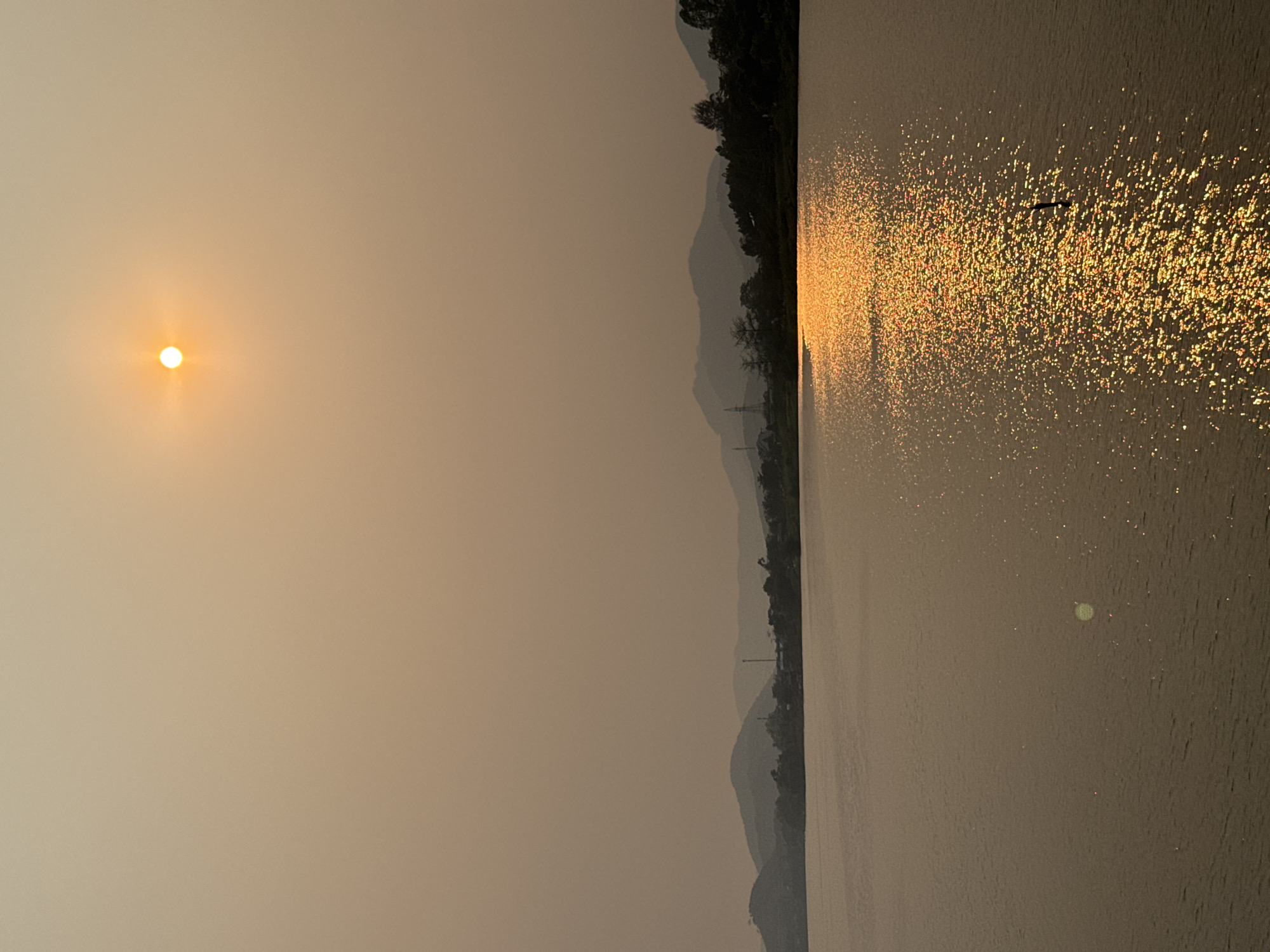 The image captures a sunset scene over a body of water in Kim Long Ward, Vietnam. The sun is positioned in the upper left corner, casting a warm, golden light across the sky. In the distance, a series of mountains are visible, partially obscured by haze. The water reflects the sunlight, creating a shimmering effect. A small bird can be seen flying across the reflection. The overall environment suggests a tranquil and hazy atmosphere.