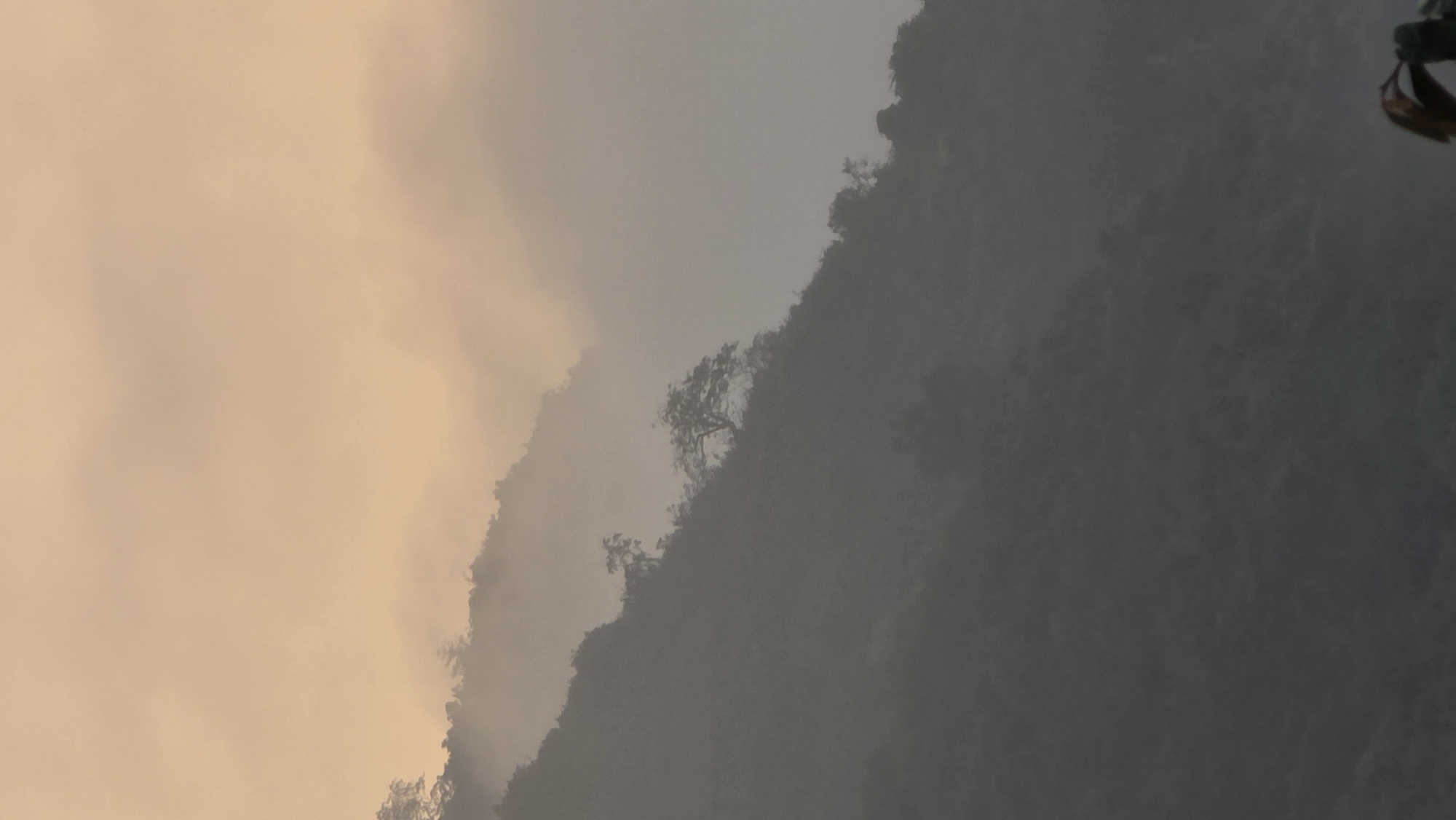 The image shows a mountainous landscape in the Bac Natural Heritage Area Trang, Vietnam. The scene is dominated by a mountainside covered in trees. The sky is filled with a thick, hazy mist, with a golden hue. The combination of fog and sunlight creates a subdued and diffused light across the scene.