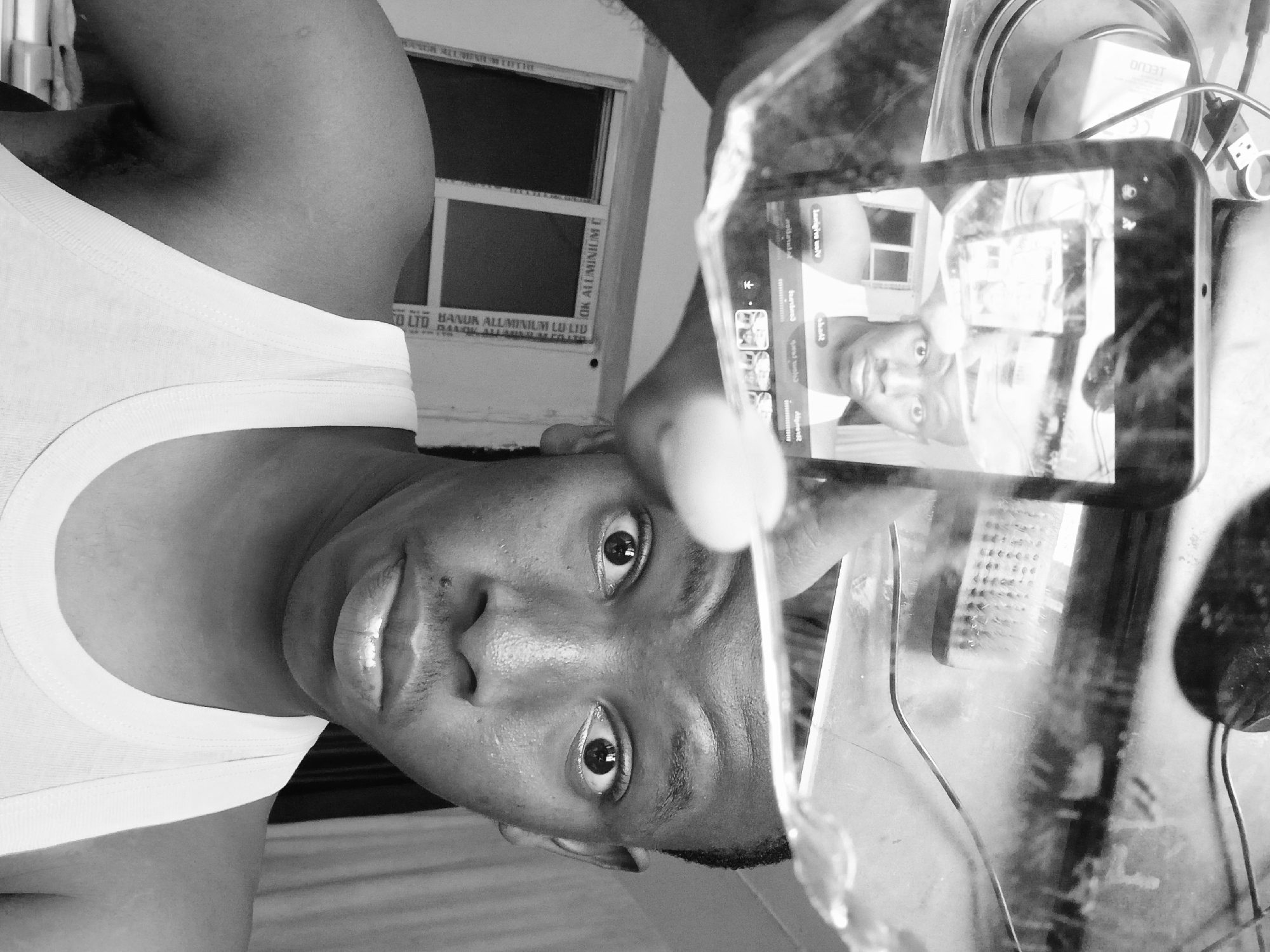 A monochrome, close-up image shows a dark-skinned individual wearing a white tank top, looking directly at the camera. The person is holding a smartphone or tablet in their right hand, displaying a mirrored image of themselves. The reflection shows a window and a light-colored wall. Part of the window frame has the words “CO LTD BANUK ALUMINIUM LU LID” and “OK ALUMINIUM C” written on it. The camera is also capturing other objects, including a charger labeled “LECUO” and a container. The overall setting suggests an indoor environment. The image’s orientation appears to be rotated counterclockwise. Although the location is Nigeria, the specific location in the city of Lagos cannot be ascertained from the details shown.