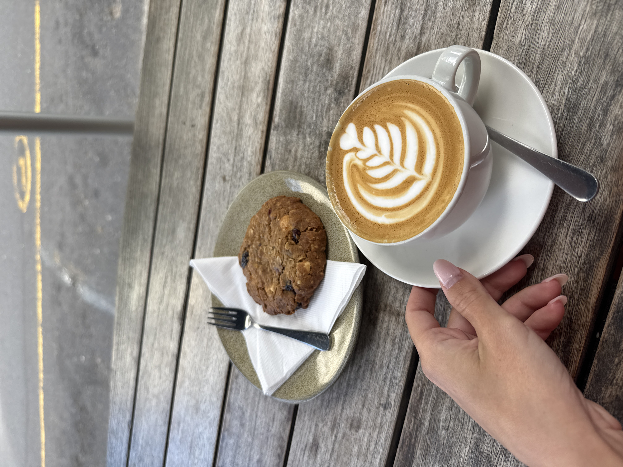 The image depicts an outdoor scene at a cafe, likely in Cape Town, South Africa, given the light and architectural style. A cup of latte with latte art rests on a white saucer, a small silver spoon laying next to it. A cookie sits on a small plate with a fork on top of a white napkin. A woman's hand is holding the coffee cup. The setting is a weathered wooden table with parallel planks. In the background, part of a gray sidewalk and metal railing are visible.