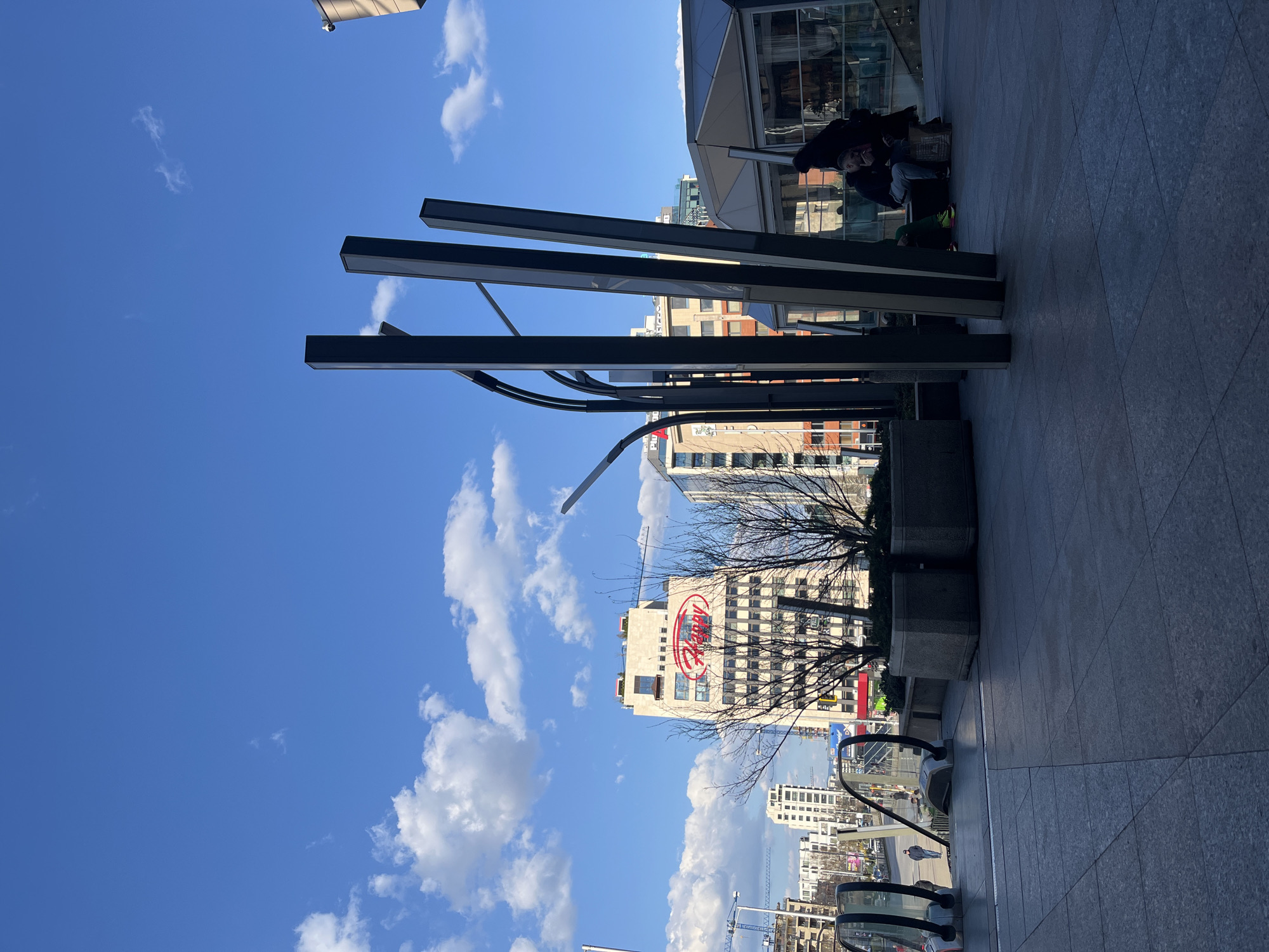 The image shows a street scene in Sofia, Bulgaria. In the background, there is a tall building with a prominent red logo, identified as the Happy Bar & Grill building. The sky is blue with scattered white clouds. In the foreground, there is a modern architectural structure with black rectangular bars. There are also escalators leading down to a lower level. A person wearing dark clothing and a mask is sitting on the ground near a building entrance. The scene is captured from a slightly elevated perspective.