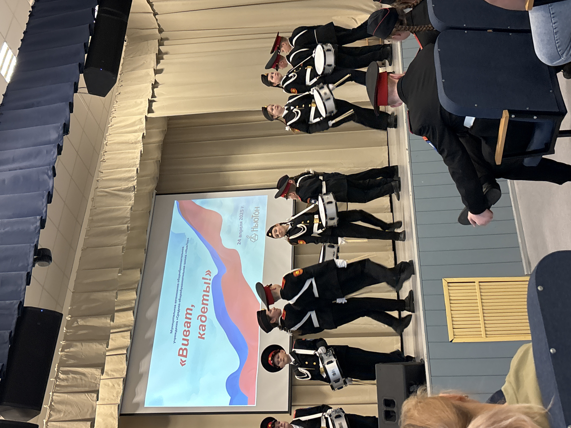 This image captures an indoor event, likely a performance or presentation, taking place on a stage within an assembly hall or auditorium in Chaikovsky, Russia.
The primary subjects are a group of approximately 6-7 young people, appearing to be cadets, dressed in formal black uniforms with white trim, black hats (some with red bands or visors), and black shoes. Each cadet holds a small, white marching drum with straps. They are arranged in rows, standing upright and appearing focused, suggesting a ceremonial or musical performance.
To the right, an audience is partially visible, seated in blue upholstered chairs. The back of a person's head with blonde hair is prominent in the foreground, indicating an audience perspective.
Behind the cadets, a large projector screen displays text and a graphic. The visible text, from the OCR, reads: "Муниципальное автономное общеобразовательное учреждение «Средняя общеобразовательная школа «Ньютон»" (Municipal autonomous educational institution "Secondary comprehensive school "Newton"), "«Виват, кадеты!»" (Vivat, cadets!), and "24 апреля 2025 г." (April 24, 2025). The screen also features a stylized graphic resembling a flag or ribbon in red, blue, and white colors, and the word "Ньютон" (Newton).
The setting includes a stage area with a light-colored backdrop. Above, large black speakers are mounted, and the ceiling edges are adorned with pleated fabric drapery in shades of blue and off-white/cream. A yellow slatted panel is visible on a distant wall. The overall lighting is bright and artificial. The event is clearly organized by or for the "Newton" school.