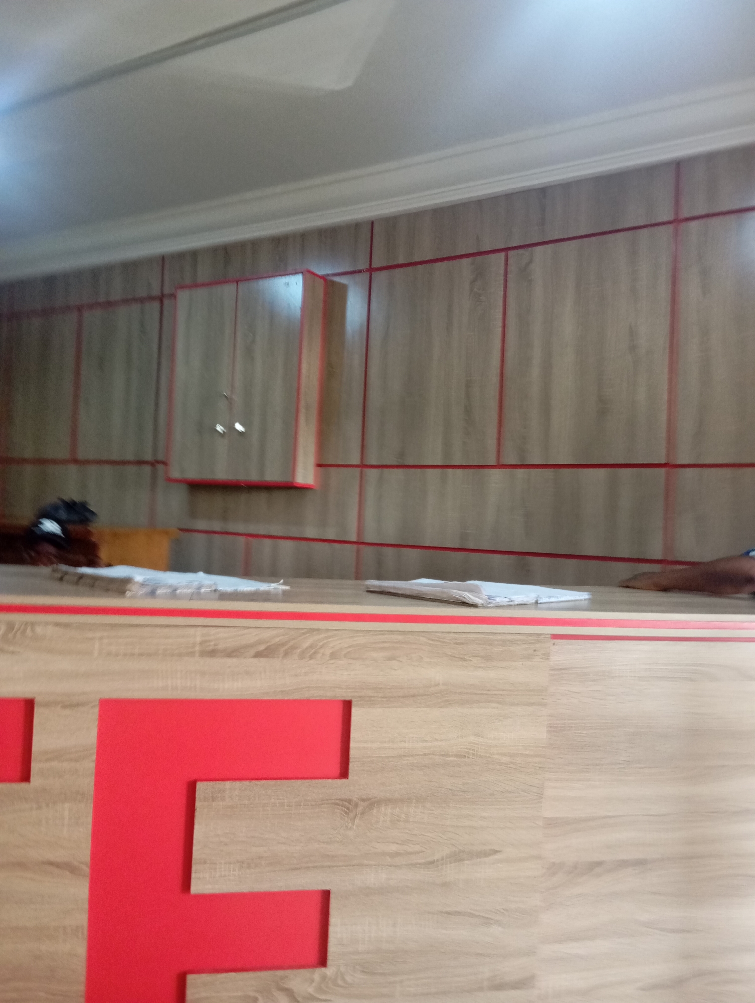 The image depicts an interior scene, possibly a reception area or office space. The walls are clad in wood-effect panels with thin red horizontal and vertical dividers, creating a grid-like pattern. Mounted on the wall is a light-colored cabinet with two doors, each with a small round handle. In the foreground, a light-colored counter with a red trim is visible. On the counter, there are stacks of white papers or documents. To the left of the counter, large red cut-out letters, the first two appearing to be "T" and "E" (or similar), are mounted on the wood-effect wall. A dark object, possibly a person or a large bag, is partially visible on the left side of the counter. The overall impression is of a modern, clean, and organized space. The exact location in Jos, Nigeria cannot be determined from the visual information alone.