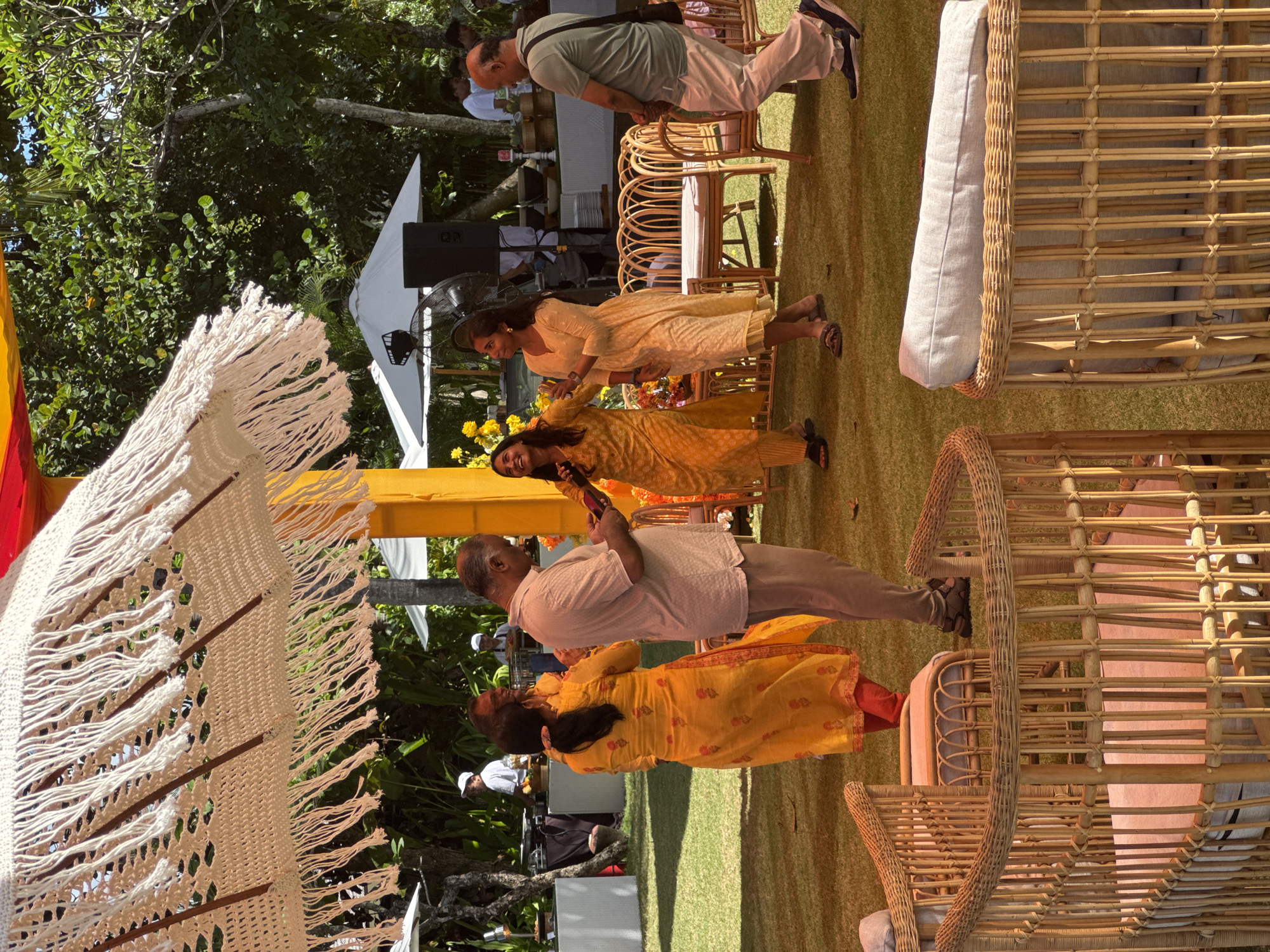 The image captures an outdoor event in Kuta Selatan, Indonesia, likely a wedding or festive gathering. The scene is set on a grassy lawn, with several people in attendance. Three women are wearing traditional Indian dresses, in the colors yellow and orange. One man is in a pale shirt and pants, while another wears an olive-green shirt and light khaki pants. The location is decorated with bamboo chairs with cushions, a yellow-and-orange canopy, and a white parasol with hanging fringe. In the background, a white tent with speakers indicates a stage or performance area. The day appears sunny, with dappled light falling through the surrounding trees.