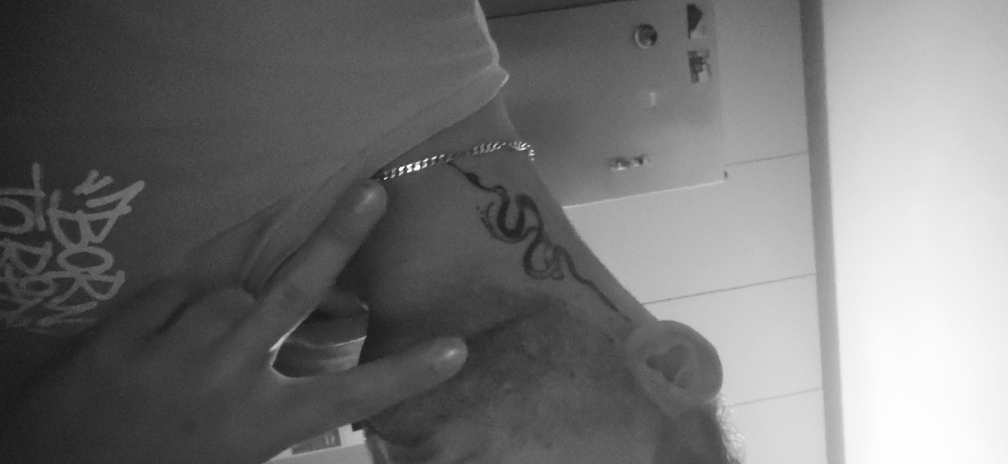 A black and white close-up photograph depicts a person's neck and shoulder. A tattoo of a stylized serpent is visible on the neck, extending from behind the ear. A silver chain adorns the upper part of the neck. A hand with two fingers extended is positioned in front of the neck, partially obscuring the tattoo. The person is wearing a t-shirt with the words "Born To" visible in a graphic design. The person's ear is visible. The background includes a light-colored wall with a light switch and other unidentified fixtures. The location is not discernible from the image.