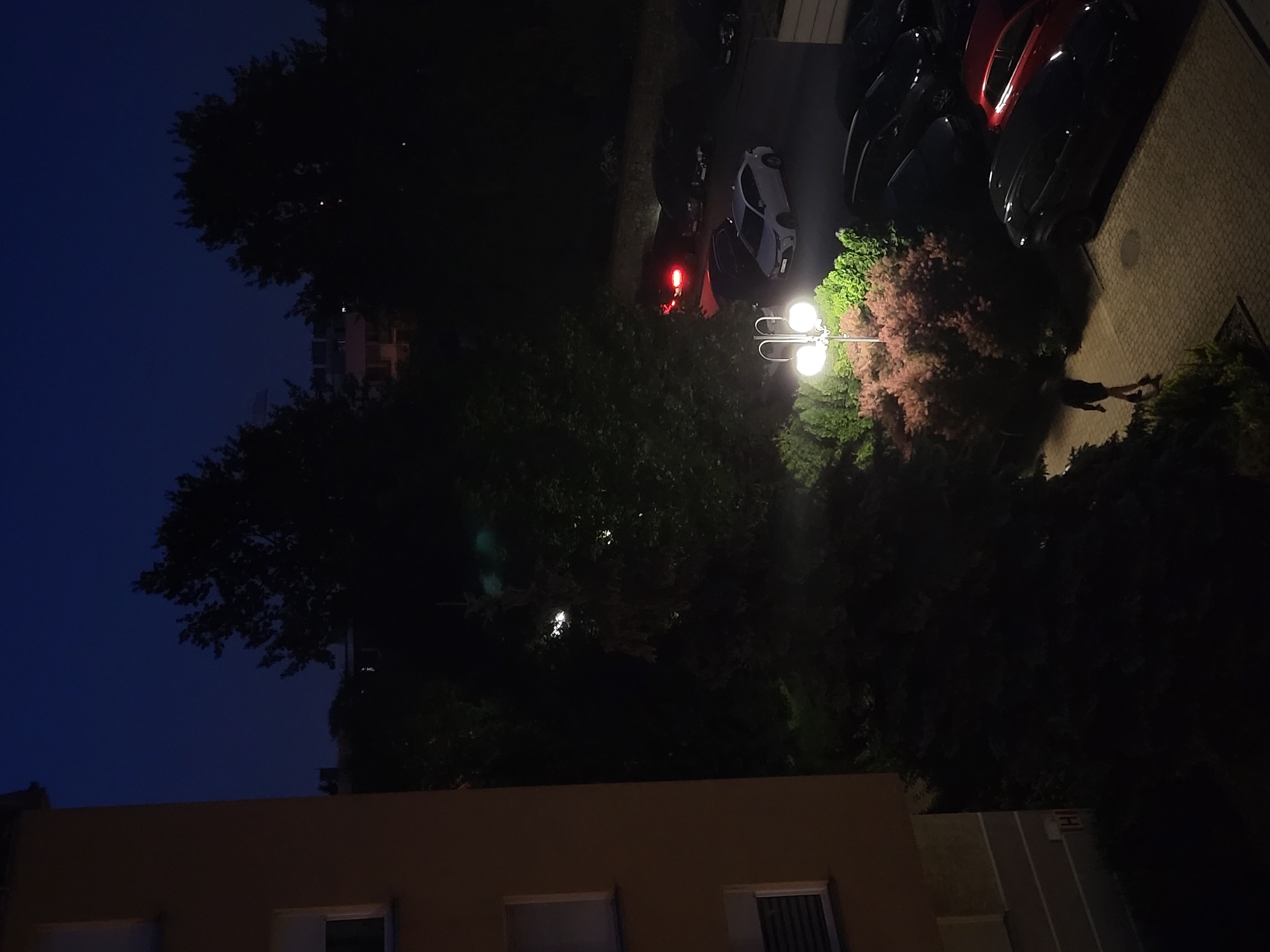 An exterior, medium-wide shot captured at night in an urban setting shows a paved area with several cars parked. A single person, silhouetted in dark clothing, walks along the pavement. To the left, a building with orange siding and white-trimmed windows is visible. The scene is illuminated by a street lamp with two globes situated in front of lush green and reddish-pink bushes. In the background, trees and other buildings are faintly discernible in the dark.