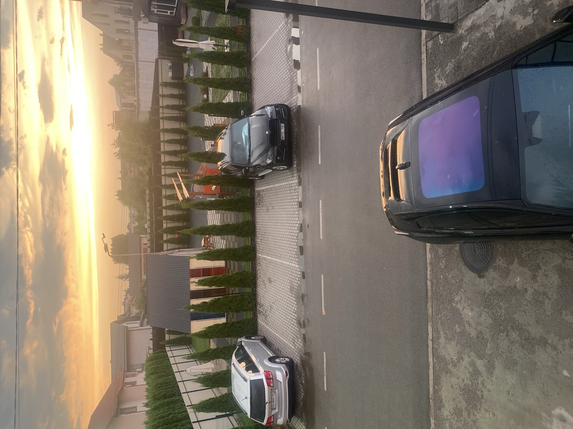 The image depicts a residential area in Novi Petrivtsi, Ukraine, during sunset. In the foreground, the roof of a black car with a panoramic sunroof is visible, reflecting the colorful sky. A wet asphalt road separates the parking area from the foreground. The parking lot, paved with cobblestones, contains two cars: a silver SUV on the left, and a black BMW SUV on the right. Behind the parking lot, a row of tall, slender evergreen trees lines a fence. Beyond the trees, several houses with pitched roofs and a small colorful shed are visible. The sky is filled with dramatic clouds illuminated by the setting sun, casting a warm, orange glow over the scene. The presence of puddles on the parking lot suggests recent rainfall.
