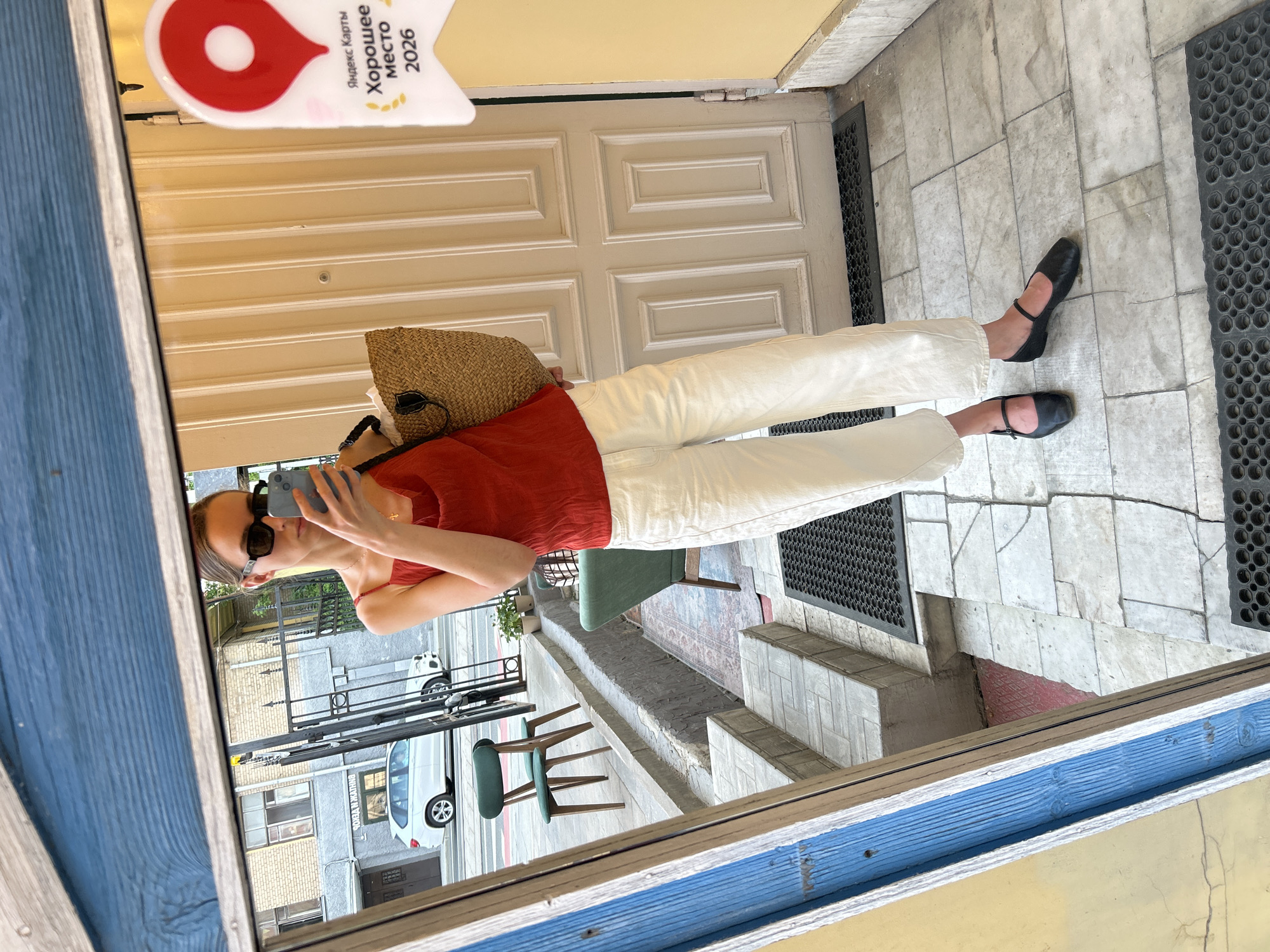 A medium shot depicts a woman taking a mirror selfie. The woman is wearing a red top, white pants, and black shoes. She is also wearing sunglasses and carrying a straw bag. The reflection shows a street scene, including a building, a car, and chairs. A sign with Cyrillic text is visible in the upper-left corner of the mirror. The location appears to be an urban area.