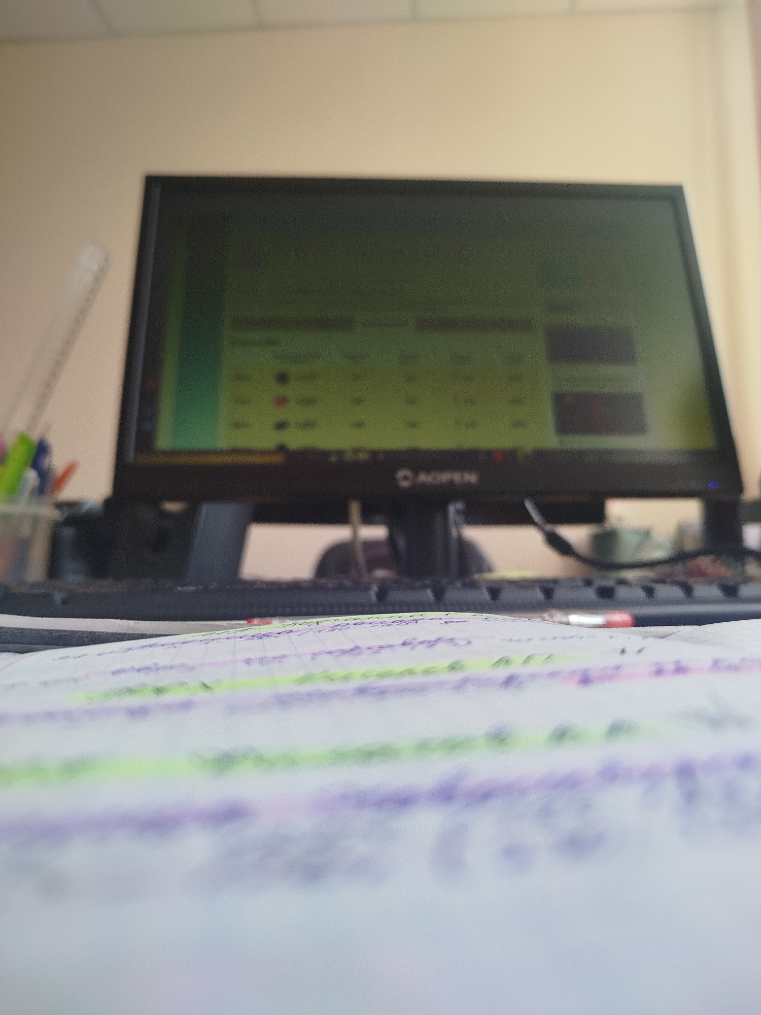 A desk scene is captured with a slightly out-of-focus computer monitor displaying what appears to be a spreadsheet or data table with green and yellow accents. The monitor has a logo that reads "AGOPEN". To the left of the monitor, a clear plastic cup holds several pens and markers in various colors, including green, blue, and orange. A clear ruler with black markings is also visible leaning against the monitor stand. In the foreground, a notebook lies open, with handwritten text and lines highlighted in purple and green. The background shows a plain, light-colored wall and a ceiling with visible panels. The overall impression is of a workspace, likely an office or study area. The image does not provide any information about the location in Zavodoukovsk, Russia.
