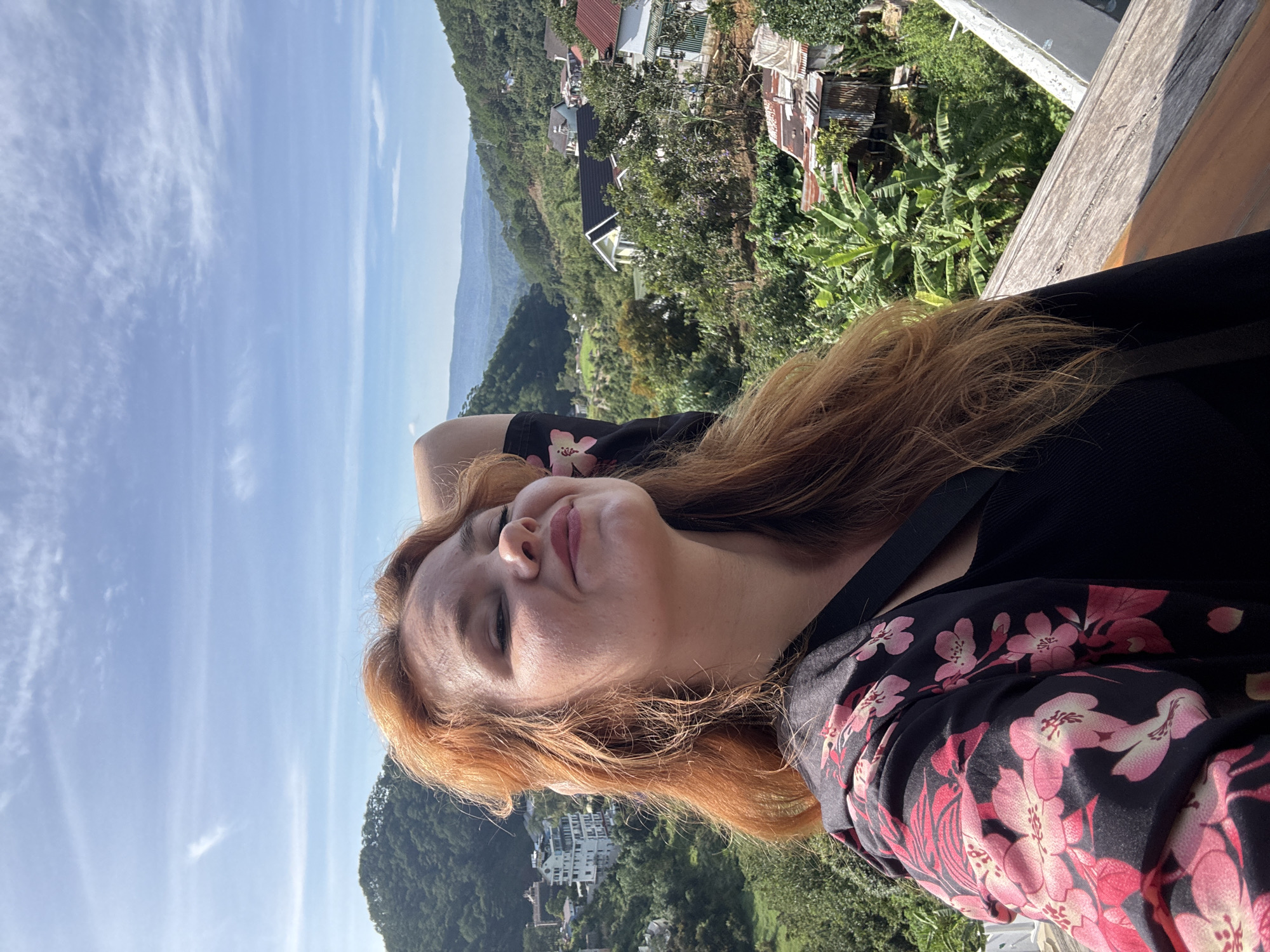 A young Caucasian woman with long, reddish-blonde hair is taking a selfie outdoors with a scenic view of a valley in Da Lat, Vietnam. She is wearing a black top and a black jacket with a pink floral pattern. Her eyes are closed, and she has a slight smile on her face as she tilts her head back. The background shows lush green hills with scattered houses and buildings, characteristic of the hilly terrain of Da Lat. The sky is blue with some wispy clouds. The woman is standing on a wooden railing, and part of a white structure is visible in the foreground.