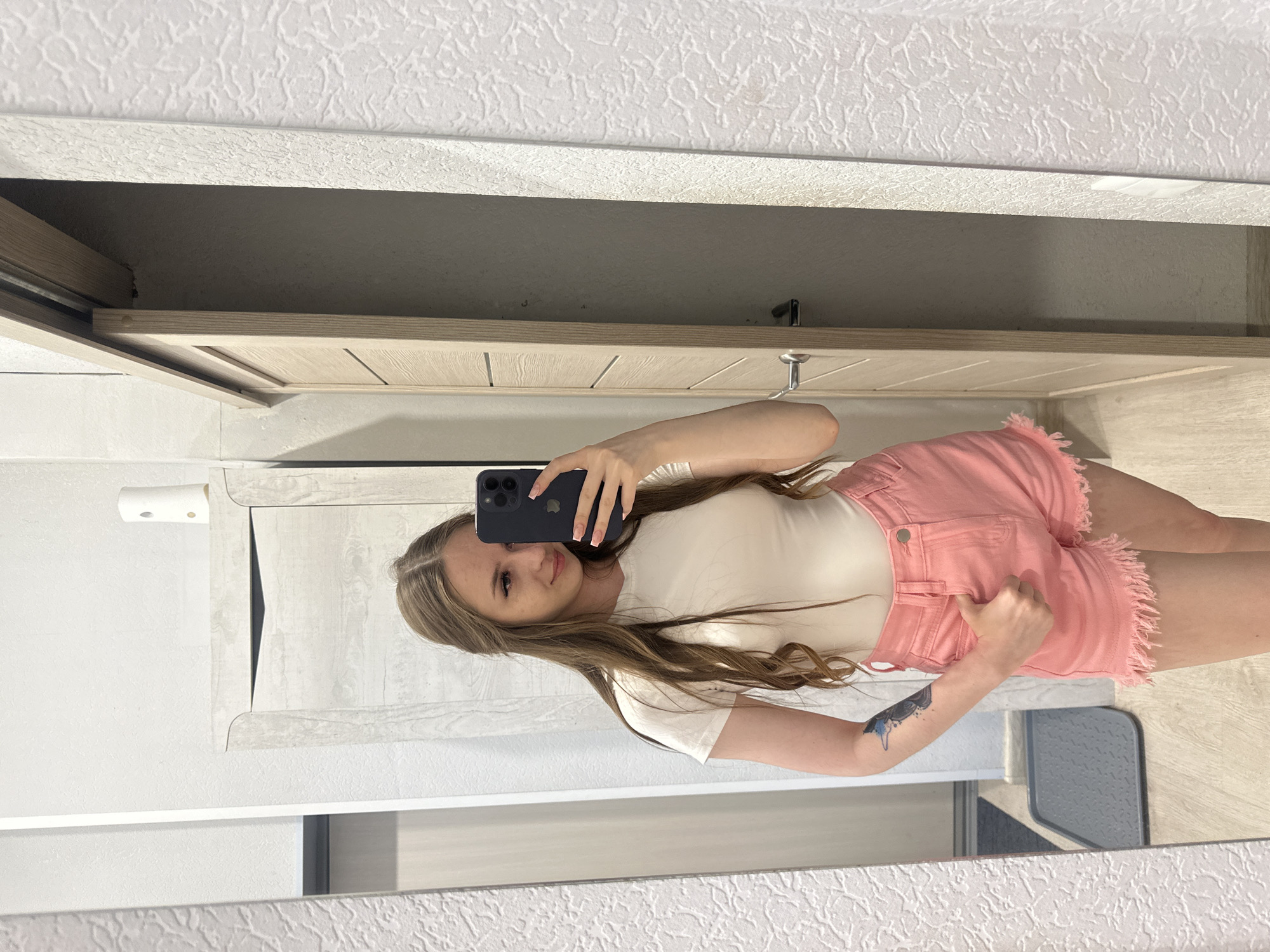 A young woman with long, light brown hair stands in front of a mirror, taking a selfie. She is wearing a white t-shirt and pink denim shorts with frayed edges. She has a tattoo on her left arm. The background shows a doorway and a patterned wall. The floor is wood-paneled, and a grey mat is visible near the doorway. A white object, possibly an air freshener, is on top of a cabinet to the left. The scene appears to be indoors, likely in a residential setting.