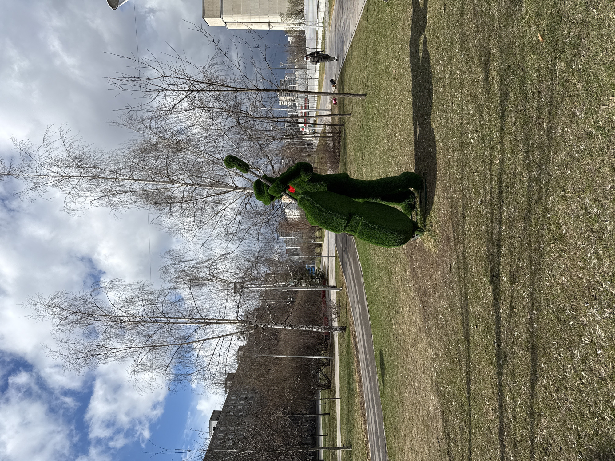 A low-angle, slightly-rotated outdoor shot depicts a green, moss-covered sculpture of a reindeer with a red nose and antlers positioned on grass adjacent to a gray concrete walkway in Moscow, Russia. The walkway is parallel to a grassy area which occupies the right of the frame. Several slender, bare trees are scattered throughout the scene, their branches reaching towards a partly cloudy sky. A tall, gray building is visible in the background, with a person walking a dog in the distance on the walkway. The composition includes elements of urban landscape and park scenery.