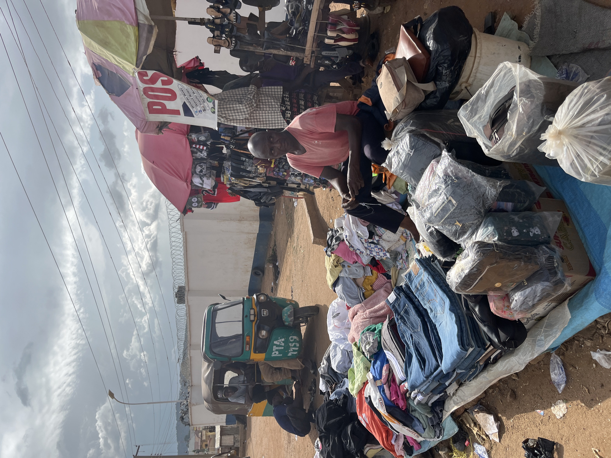 This image captures a street market scene in Jos, Nigeria. In the foreground, a vendor sits on the ground amidst a colorful display of folded clothing, including jeans, shirts, and other garments, spread out on a blue tarp. Several bags, some wrapped in plastic, are piled nearby. Behind the vendor, a green and yellow auto-rickshaw is parked, with two individuals inside and one kneeling beside it, appearing to be working on the vehicle. In the background, a white wall topped with barbed wire separates the market from other structures. Two market stalls with umbrellas are visible, one displaying clothing and accessories, and another with shoes arranged on racks. A sign on one of the stalls reads "POS" and lists various financial services. The ground is dusty, and power lines crisscross the sky, which is partly cloudy. The overall impression is of a bustling, open-air marketplace.