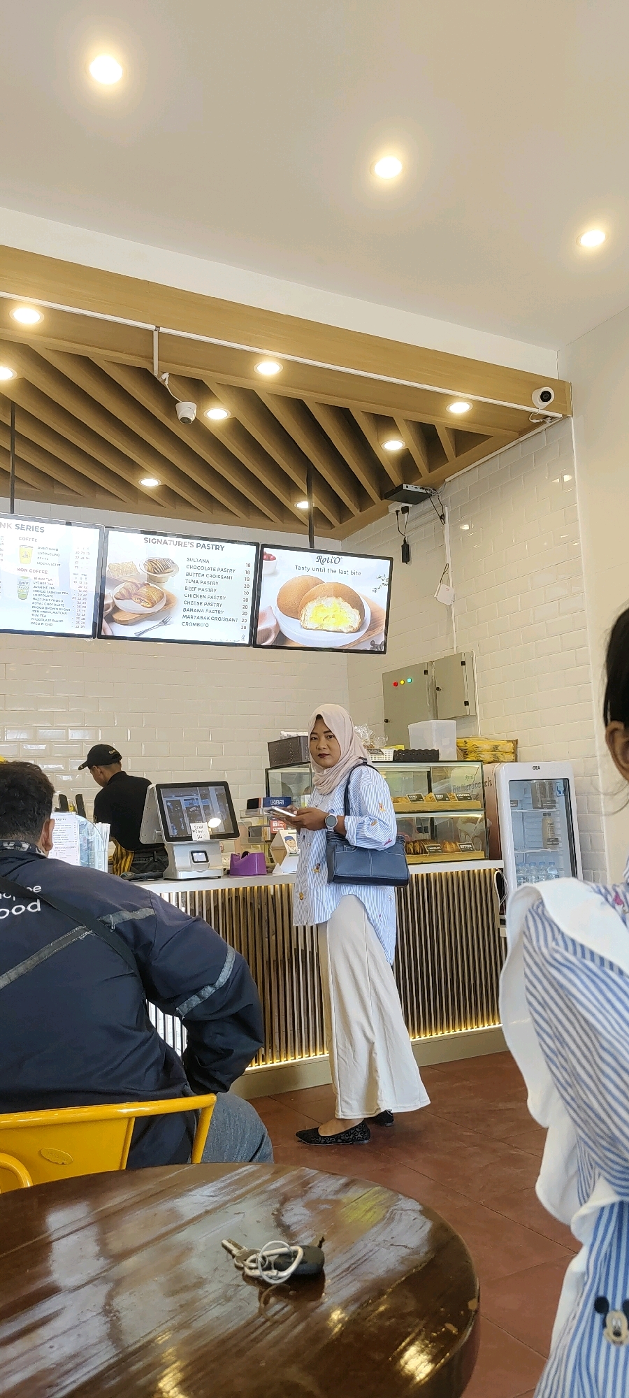 A woman wearing a hijab and a striped blue and white shirt is standing at a counter in what appears to be a cafe or bakery. She is holding a smartphone in her right hand and a dark blue handbag on her left shoulder. She is wearing light-colored wide-leg pants and black flat shoes. Behind her, a man wearing a black cap and apron is working behind the counter, which features a touchscreen ordering system. To the right of the ordering system, there are display cases showcasing pastries and a refrigerator. Above the counter, digital menus are displayed on screens. The cafe has a modern interior with wooden accents on the ceiling and white tiled walls. The floor is tiled with reddish-brown tiles. In the foreground, a round, dark wooden table is visible with a set of keys lying on it. The scene is likely in Banjarbaru, Indonesia, given the context of the menu items which include "Sultana", "Chocolate Pastry", and "Butter Croissant," which are common in Indonesian bakeries.