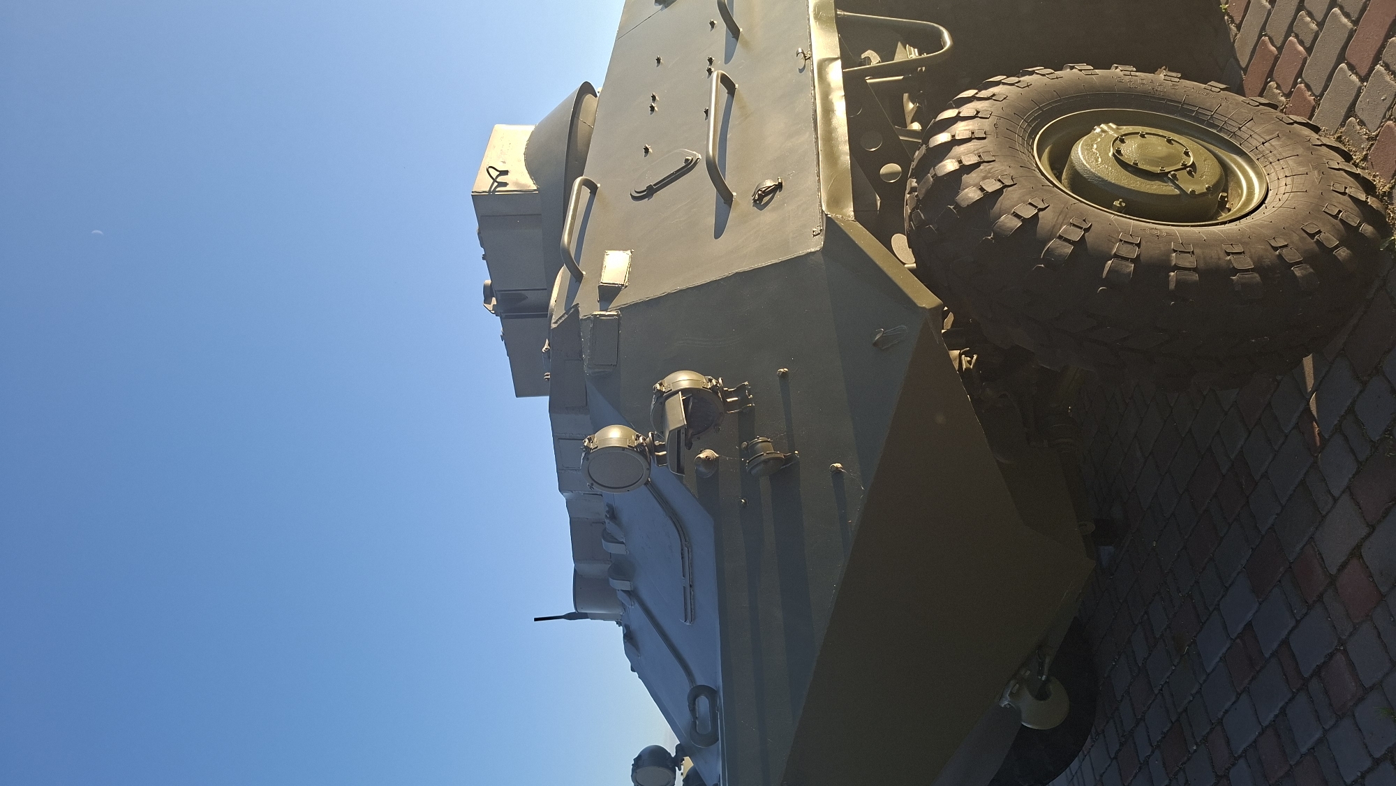 The image shows a military armored personnel carrier parked on a paved surface made of brick pavers. The vehicle is olive green and is viewed from a low angle, emphasizing its size and ruggedness. The background is a clear blue sky with a sliver of a crescent moon visible. The setting appears to be outdoors, possibly in a park or a memorial site, given the paved walkway. The lighting suggests it is either early morning or late afternoon, with the sun casting shadows. The APC has a distinctive angular design, a large tire, and several mounted lights or sensors on its turret and hull. No people are visible in the image. The location is specified as Smalyavichy, Belarus.
