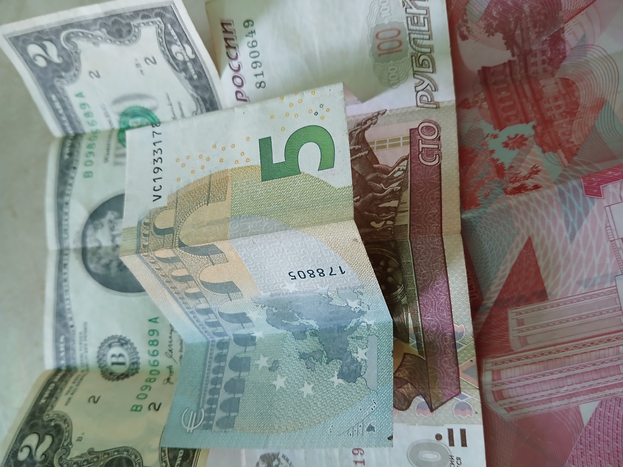 The image displays a collection of international banknotes, including a US $2 bill, a €5 Euro note, and Russian Rubles. The denominations of the Ruble notes are not fully visible. The currencies are layered and partially overlapping, indicating they have been placed together.