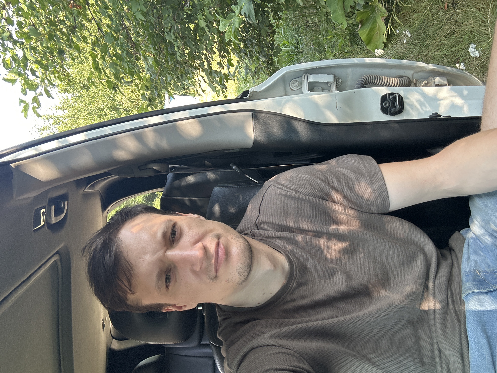 A man is sitting in the driver's seat of a vehicle, with the door open. He is wearing a brown t-shirt and blue jeans. The vehicle is parked outdoors, with trees and greenery visible in the background. The scene suggests a rural or suburban setting.
