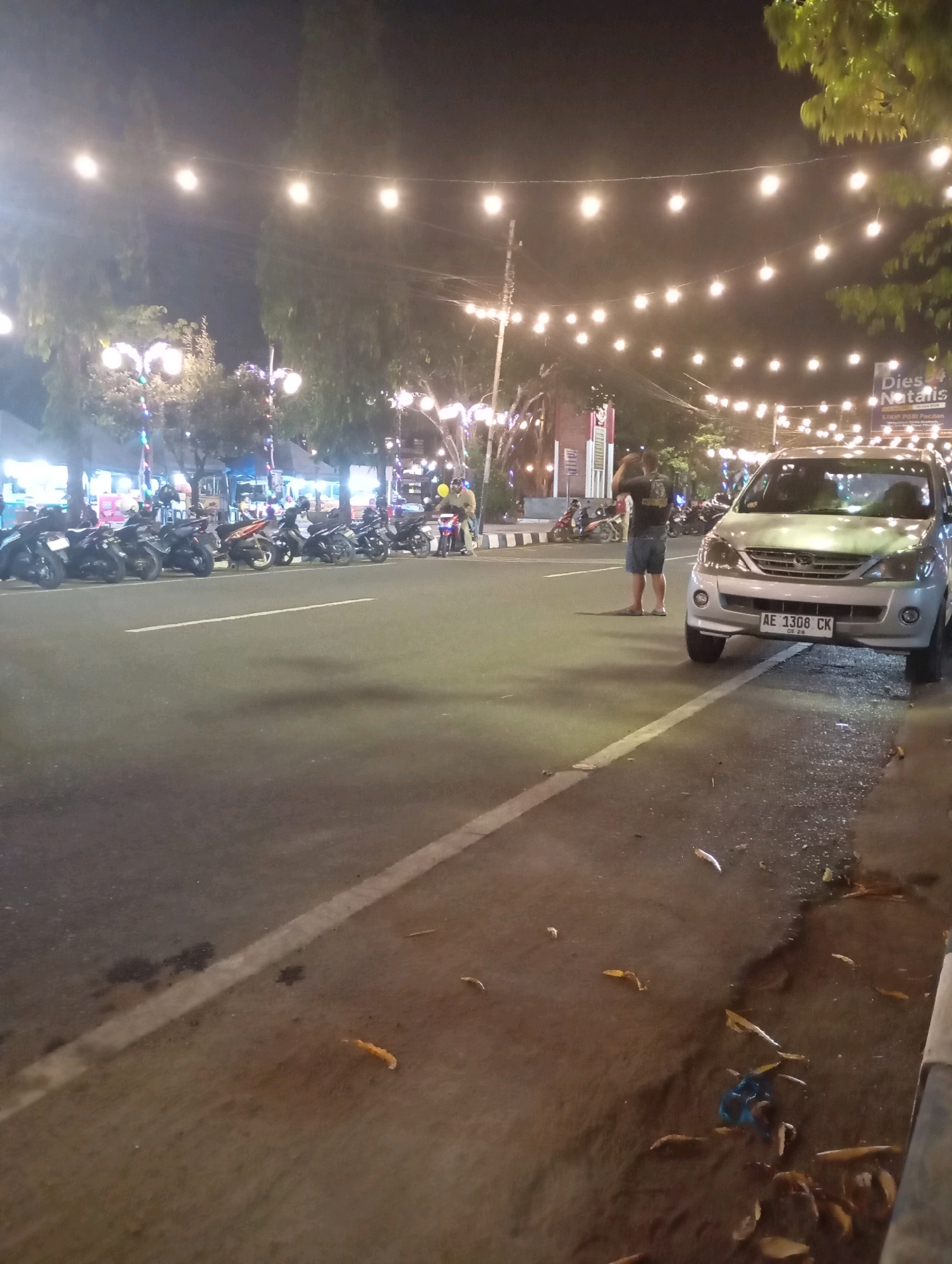 The image depicts a street scene at night in the city of Pacitan, Indonesia, likely near the city center or a public area given the presence of decorative string lights. On the left side of the image, a row of parked motorcycles lines the street. A silver minivan, with license plate AE 1308 CK, is parked in the center right of the frame. A man wearing a dark t-shirt and blue shorts is standing in the street behind the minivan, holding a phone and appearing to take a photo. In the background, several trees are visible, some with decorative lights intertwined. Buildings and awnings suggest a commercial or public establishment. The street surface appears wet in places, possibly from recent rain or street cleaning, and scattered leaves are visible on the ground. The overall atmosphere is festive due to the string lights, but the scene is also mundane, capturing a moment in everyday urban life at night.